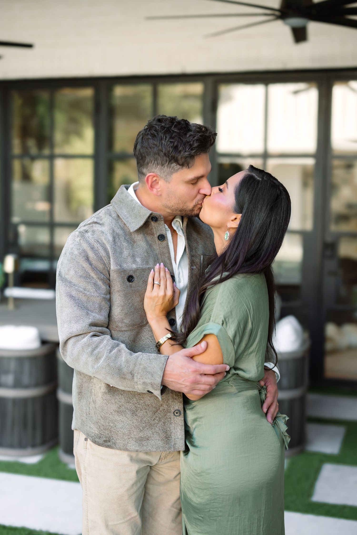 A couple shares a romantic kiss outdoors, man in gray jacket and woman in green dress, embracing on a patio.
