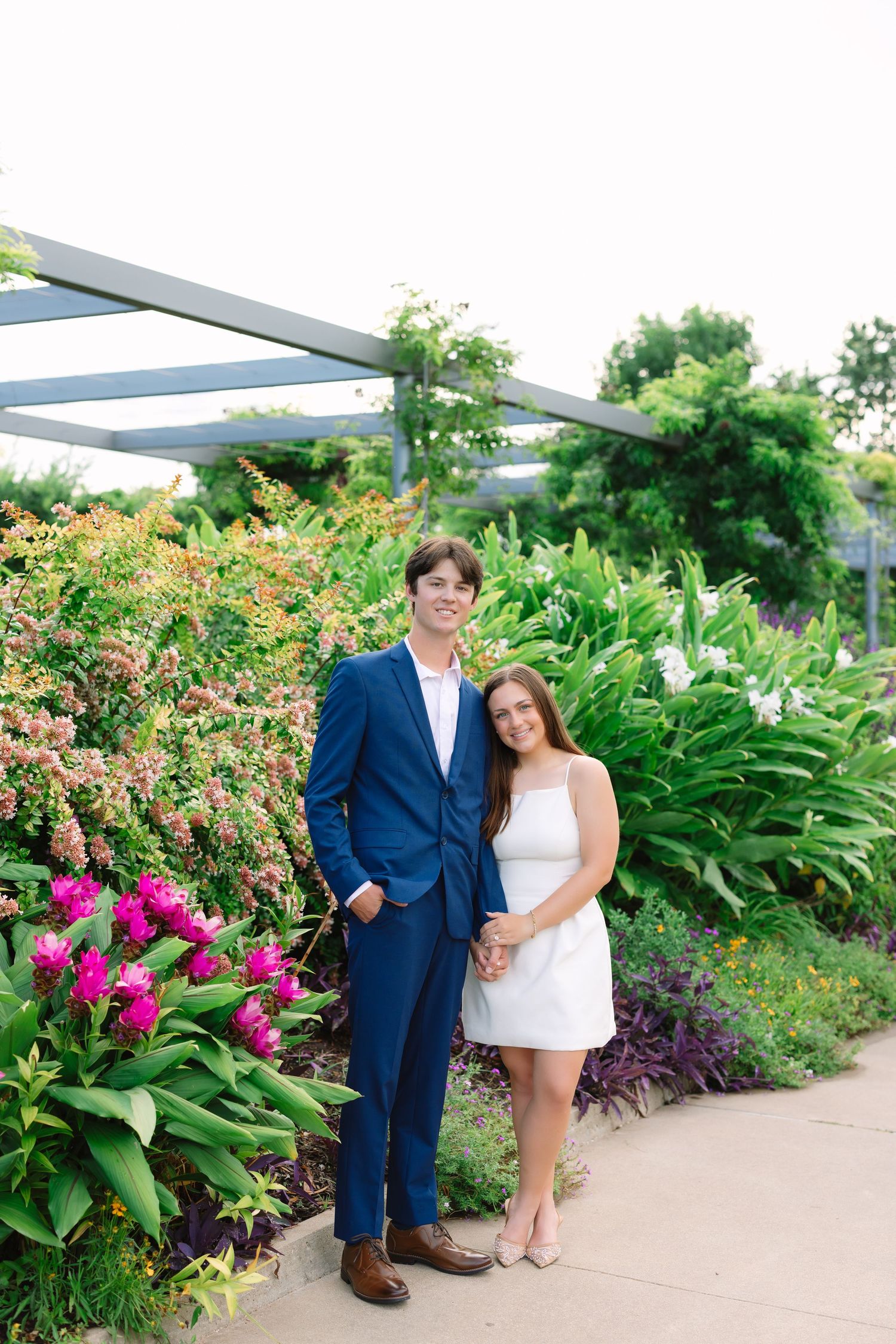 A couple poses in a garden setting with vibrant pink flowers and greenery in the background.