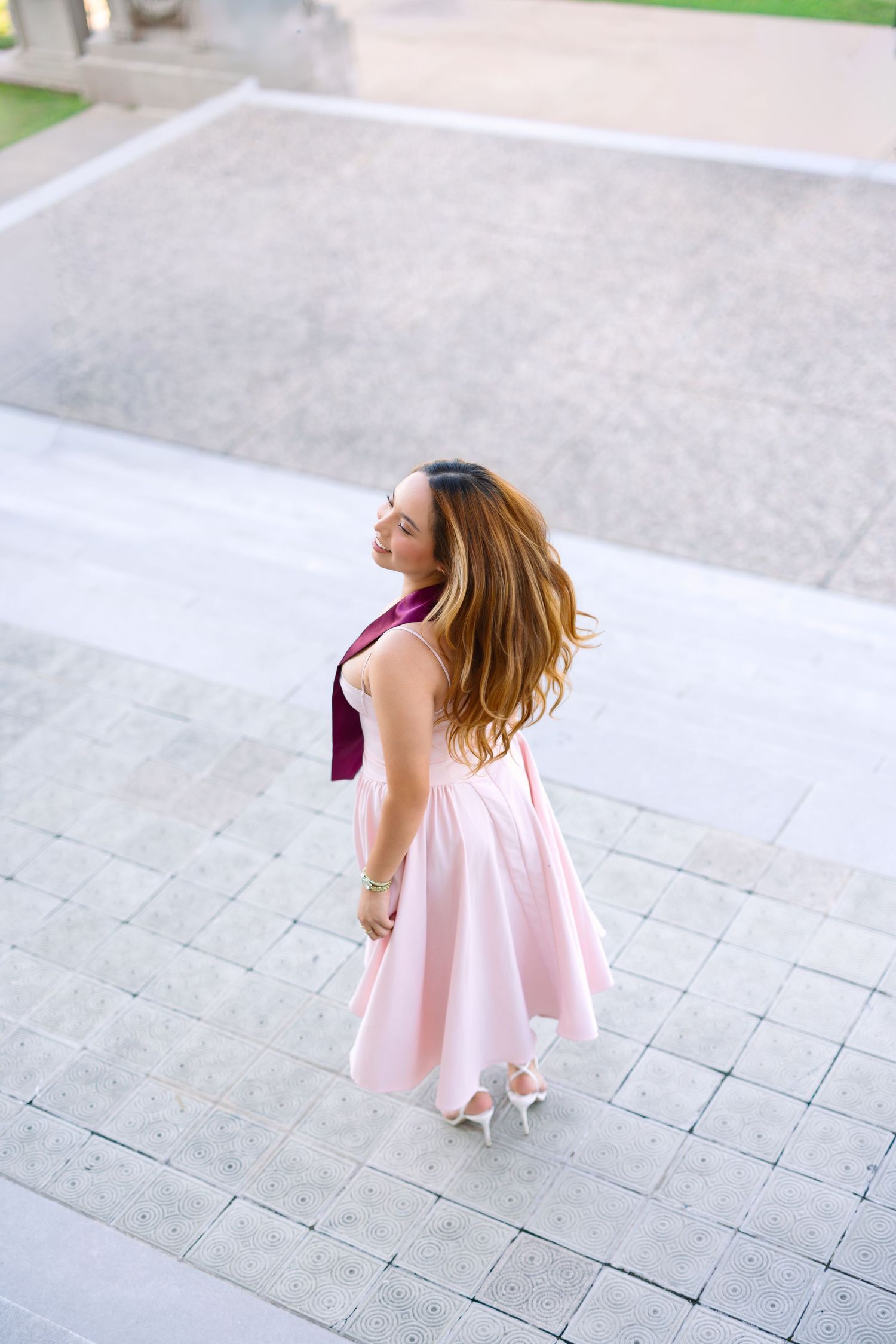 Woman in pink dress and burgundy scarf twirls on stone steps, viewed from above, hair flowing.