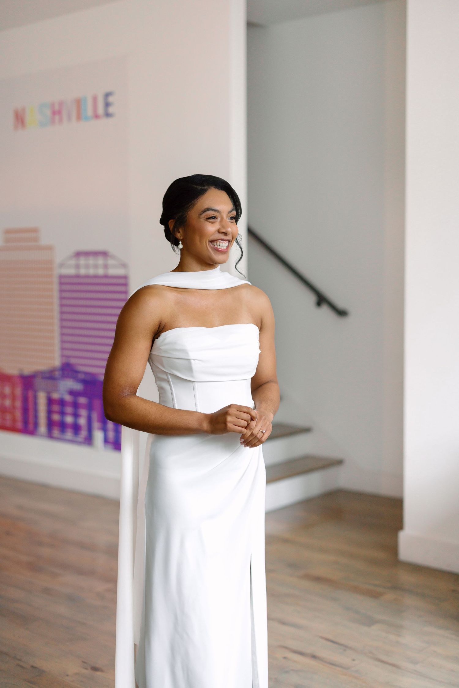 Smiling bride in white strapless gown with halter detail poses in front of Nashville skyline mural.