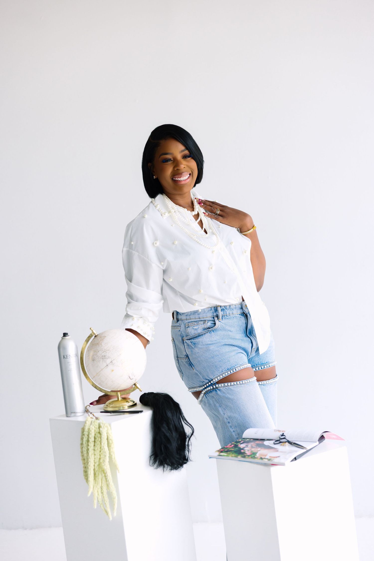 A smiling Black woman in a white shirt and ripped jeans poses beside pedestals with a globe, books, and accessories.