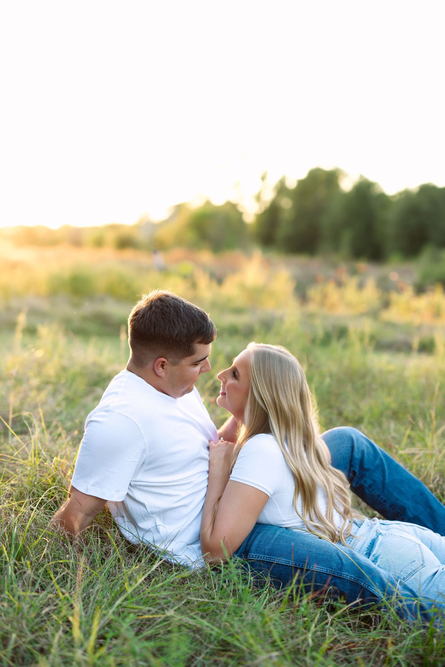A romantic couple lies in a sunlit grassy field at golden hour, gazing at each other in white shirts and jeans.