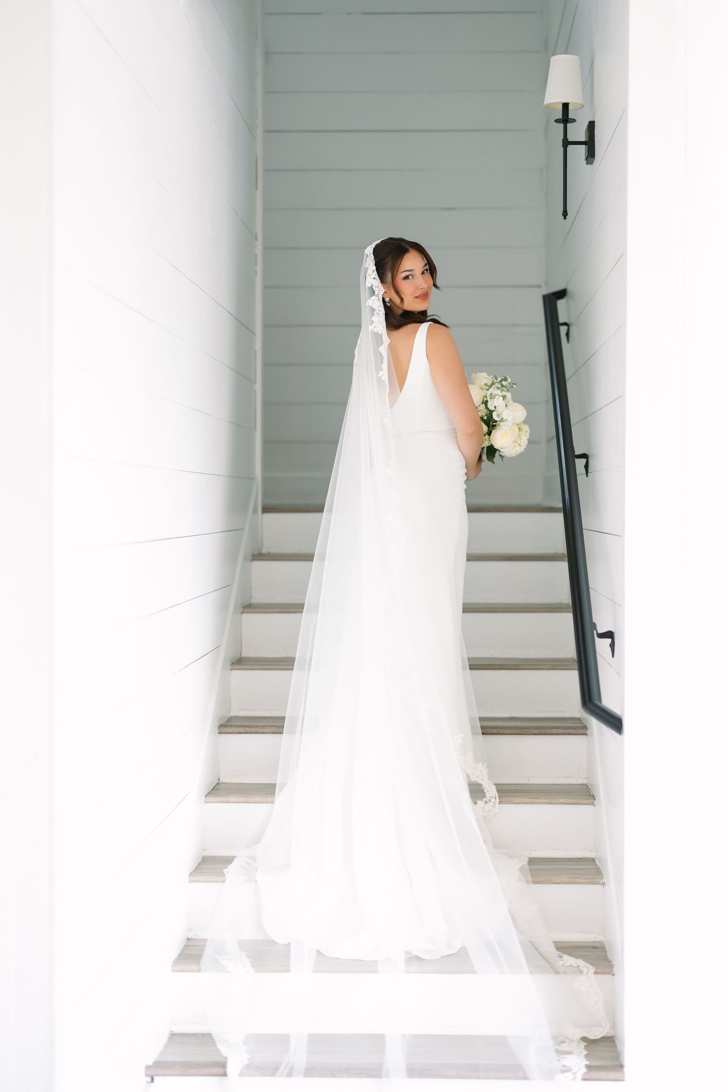 Bride in white gown and long veil holding bouquet, standing on white staircase, looking back over shoulder.