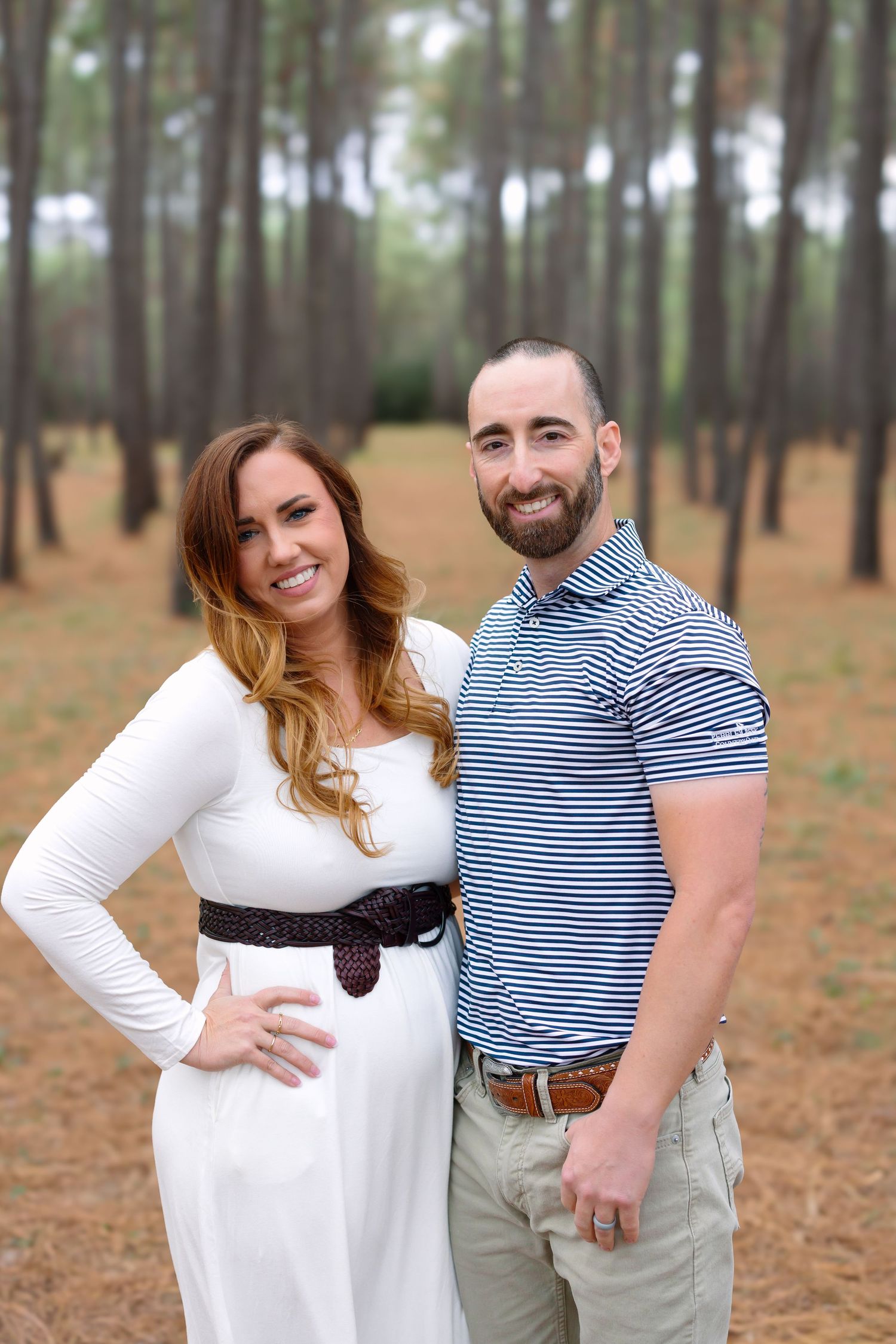 Smiling couple posing in pine forest, woman in white dress with belt, man in striped polo shirt.