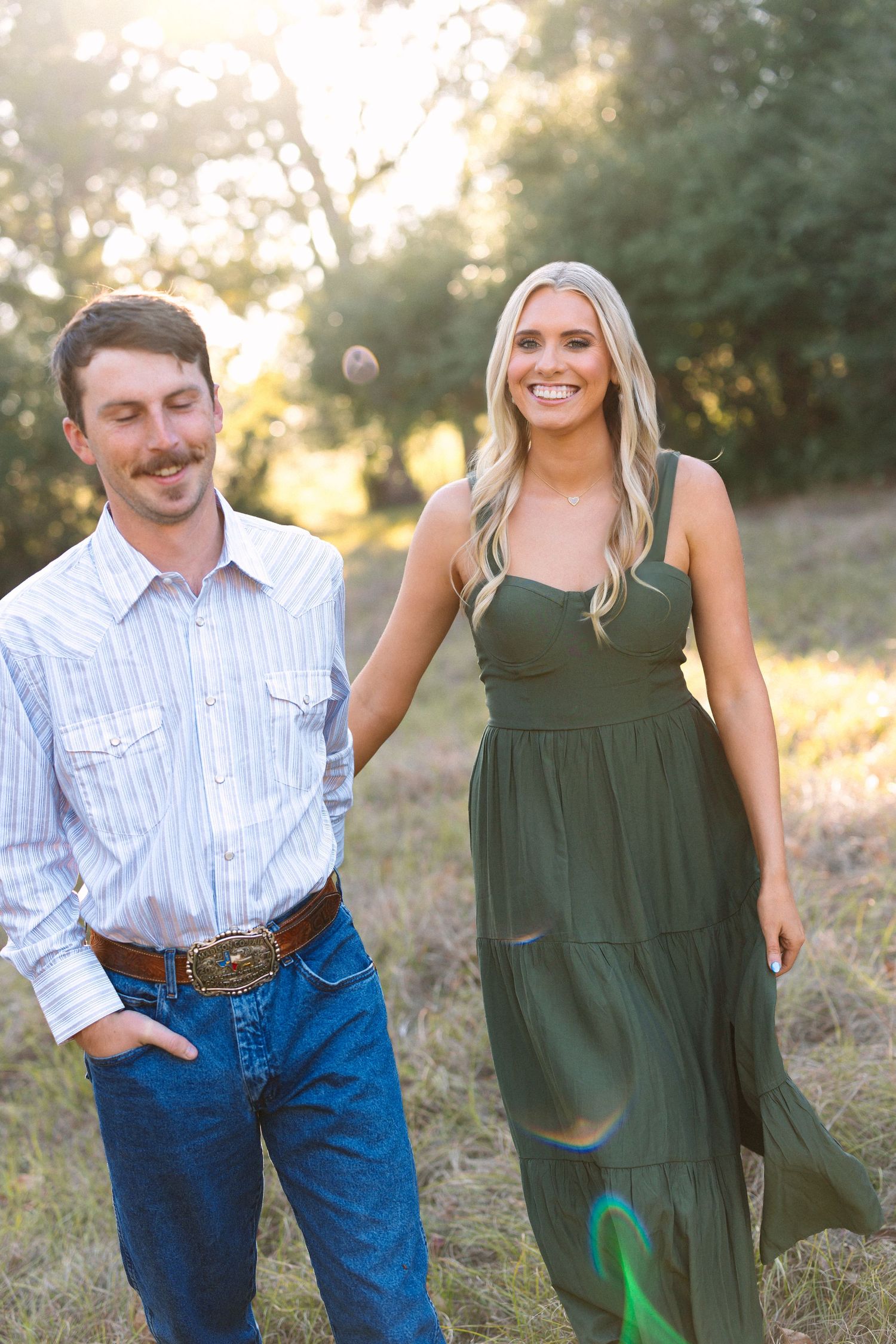 Happy couple walking in a sunny meadow, blonde woman in green maxi dress laughing with man in western attire.