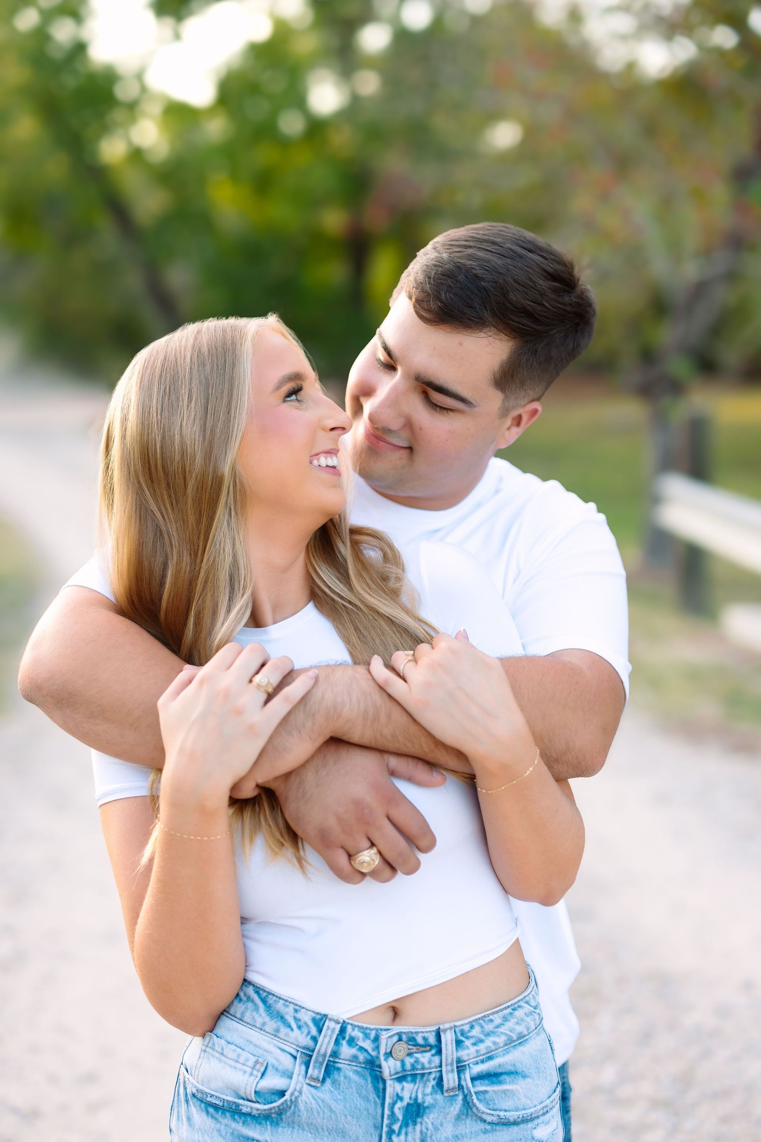 A couple embraces on a dirt path, the man hugging the smiling blonde woman from behind, both wearing white tees and jeans.