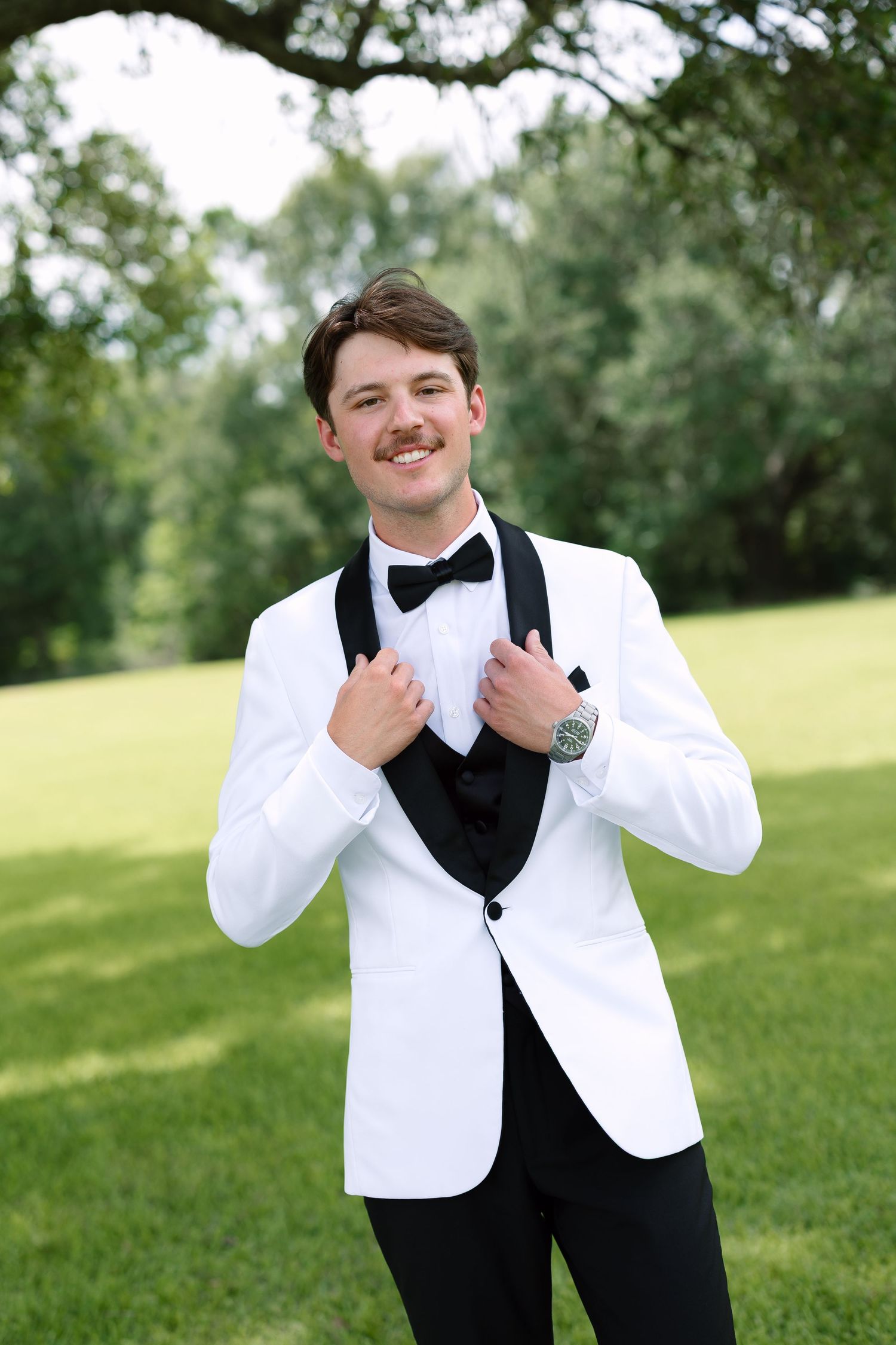 Smiling groom in white tuxedo jacket with black bow tie posing outdoors on green lawn.