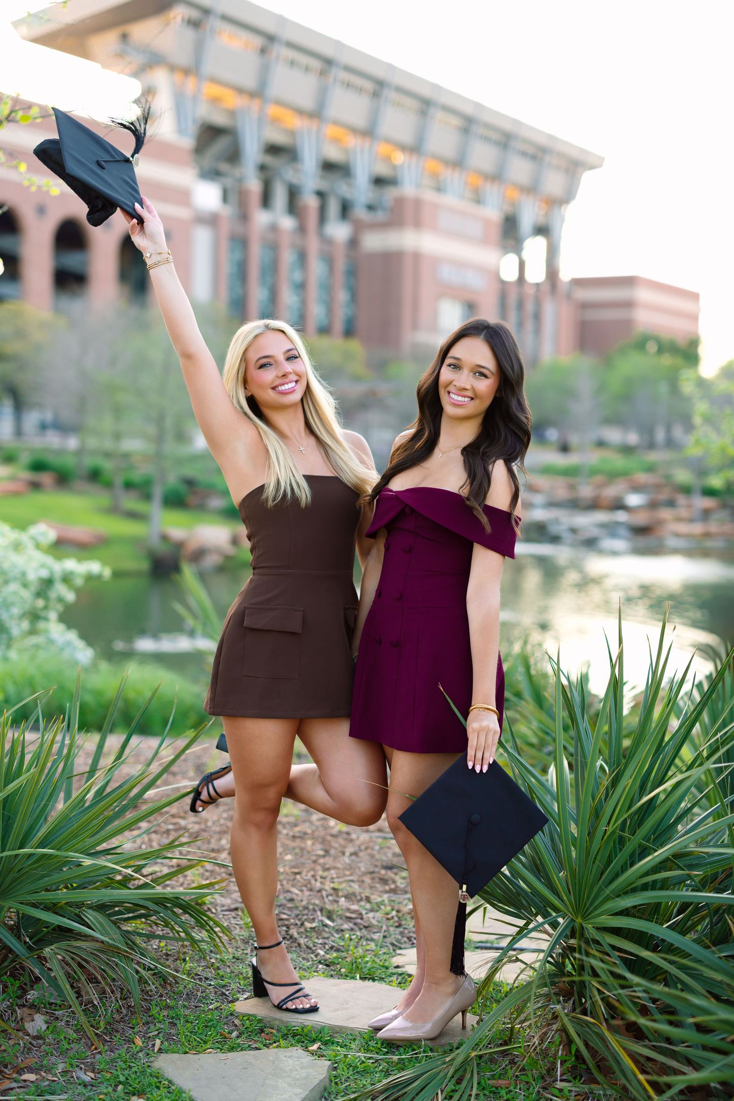 Two smiling female graduates in mini dresses celebrate outdoors, one raising her cap, near a campus pond.