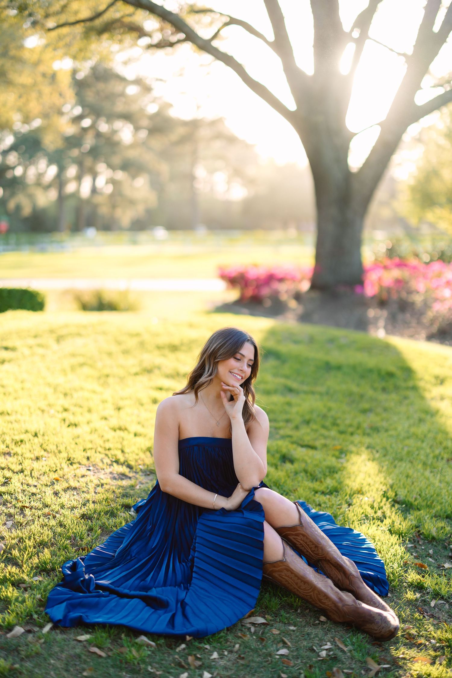 Graduate in navy dress and boots poses on sunlit grass near trees.