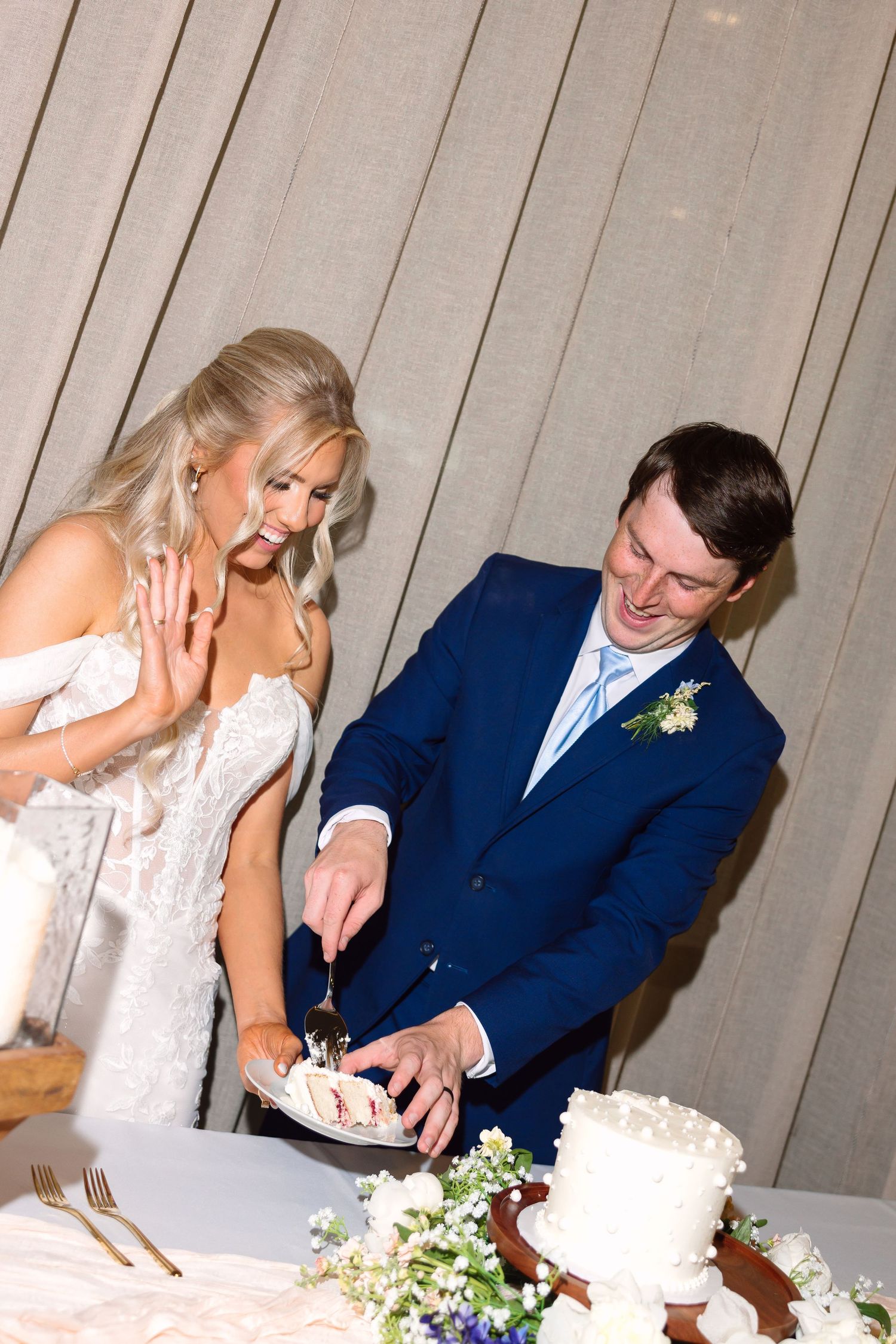 Smiling bride and groom cut wedding cake together at reception, groom in navy suit with floral boutonniere.