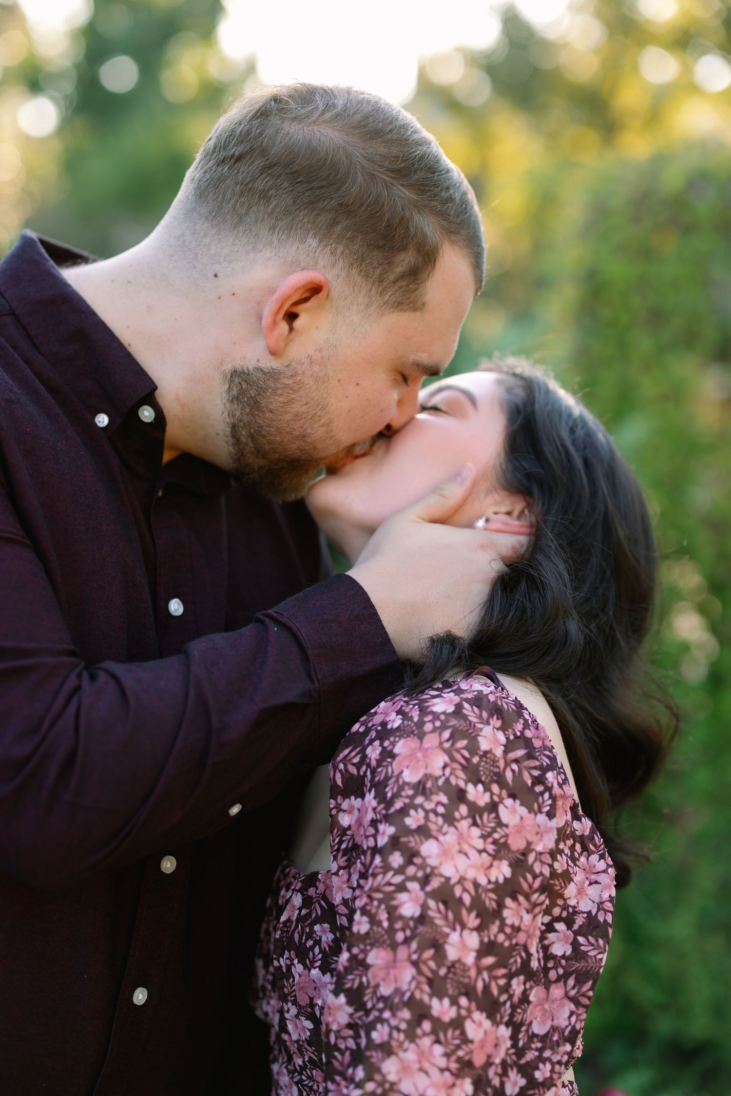 A couple shares a romantic kiss outdoors, surrounded by lush green foliage in warm golden light.