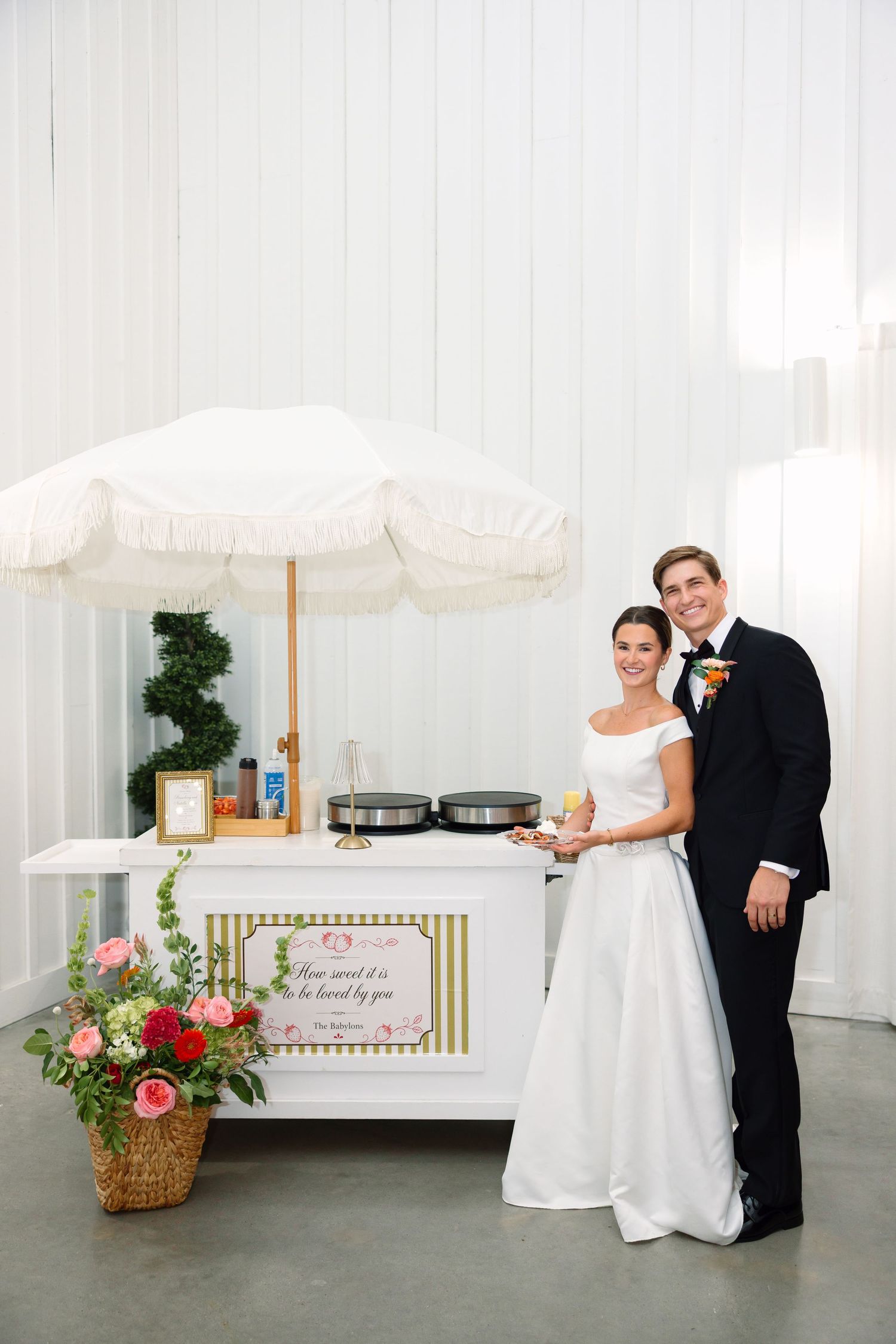 Bride and groom smiling together beside a white dessert cart with umbrella at their wedding reception.