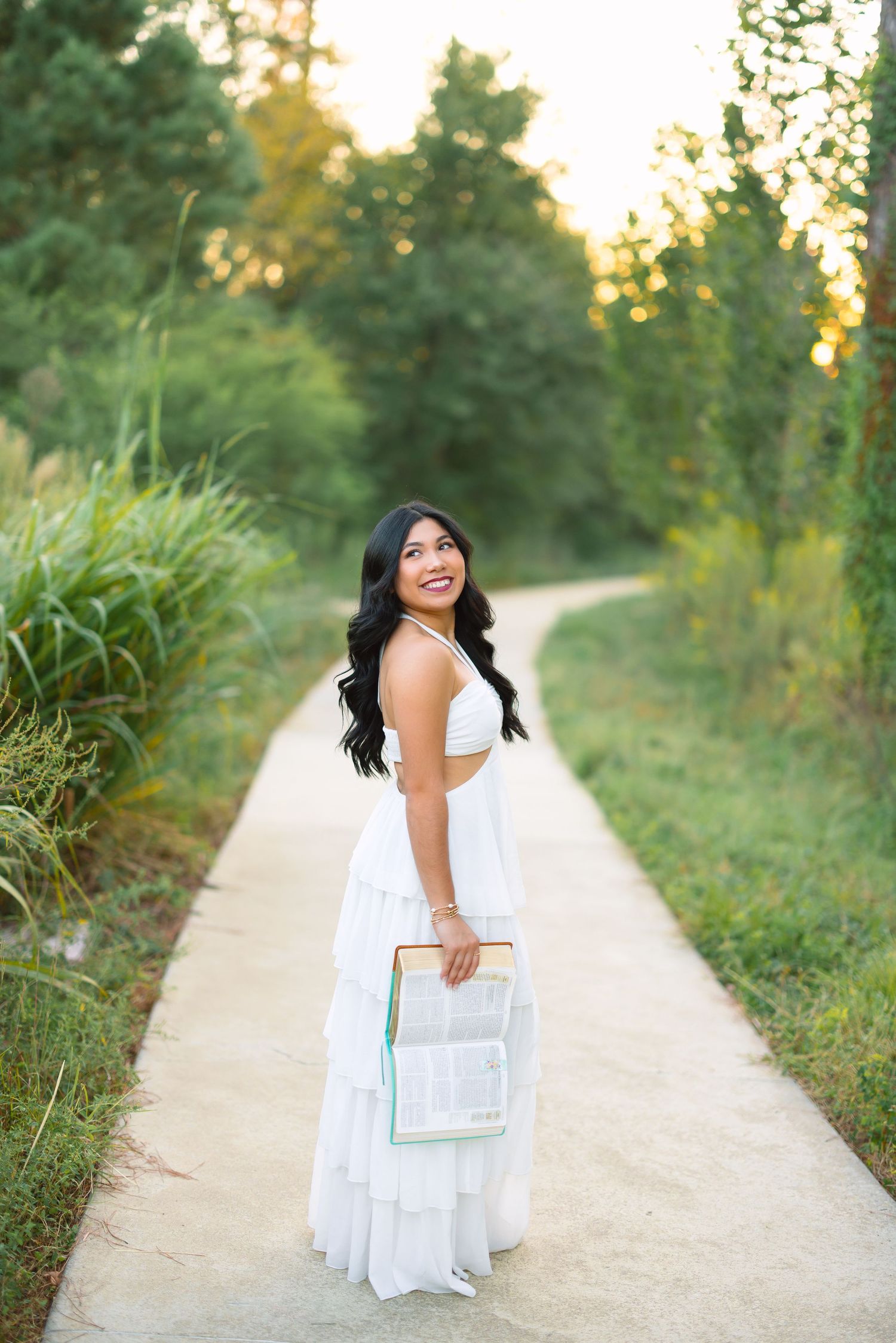 Woman in white dress holds clear bag while posing on nature path during golden hour.