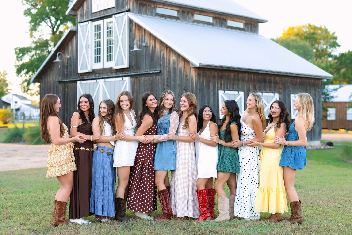 Large group in summer dresses and boots poses together in front of a rustic barn.