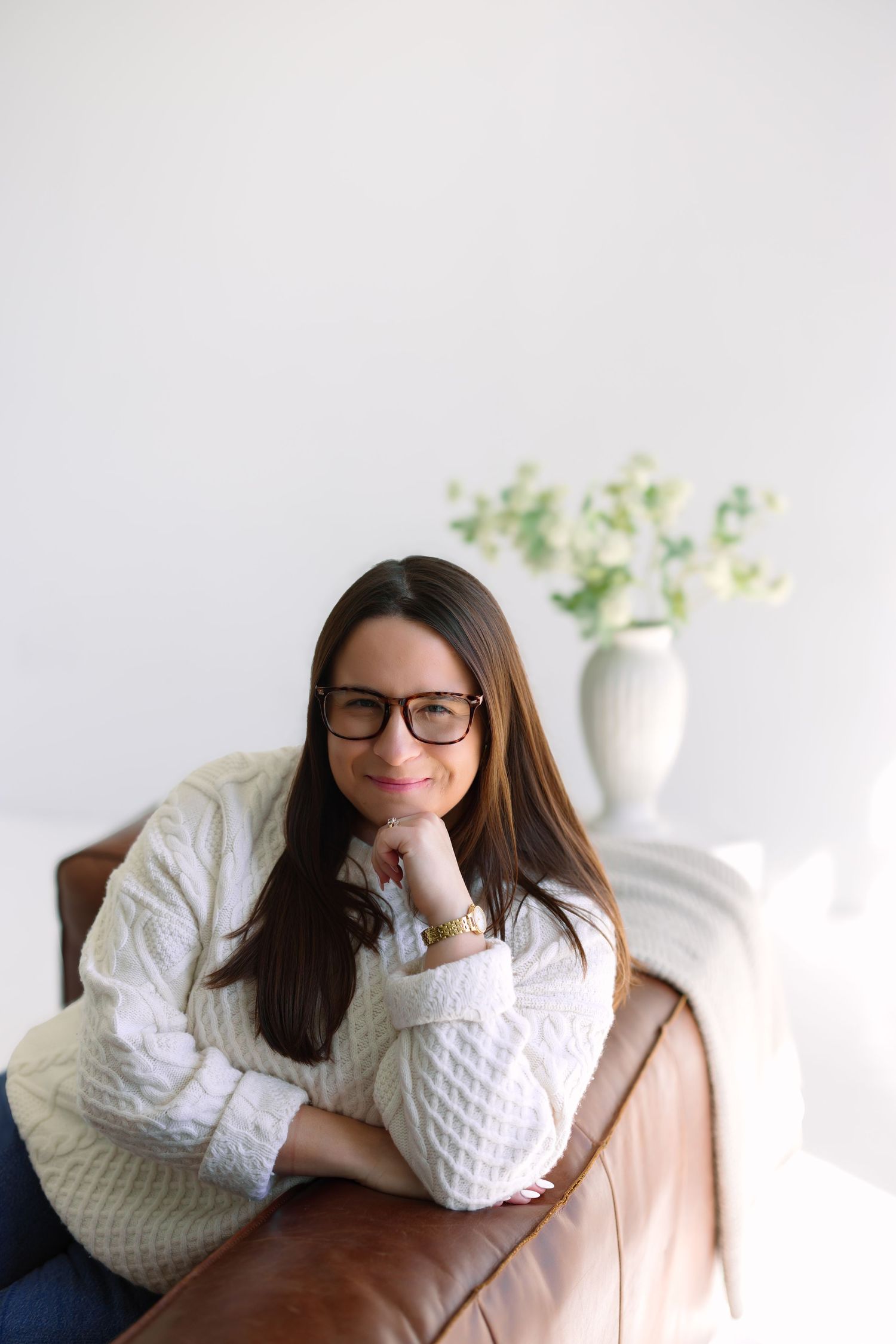Woman in white cable-knit sweater and glasses smiling while leaning on a brown leather sofa indoors.