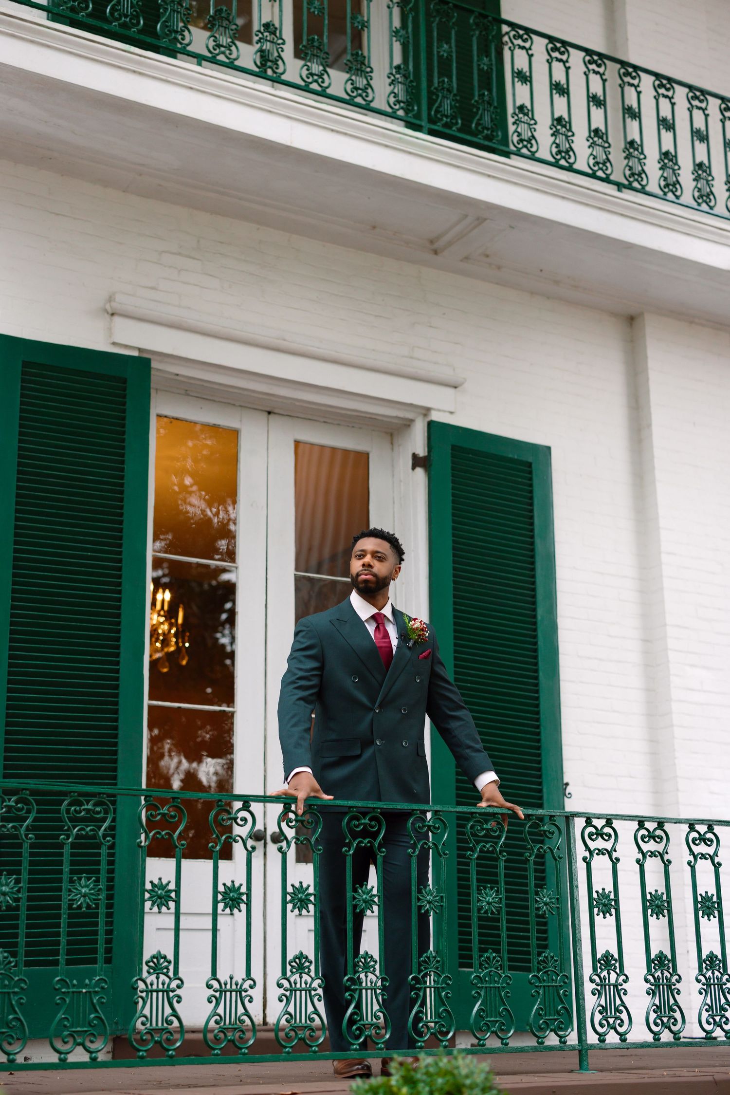 Dapper groom in dark suit with burgundy tie standing on ornate green iron balcony of white building.