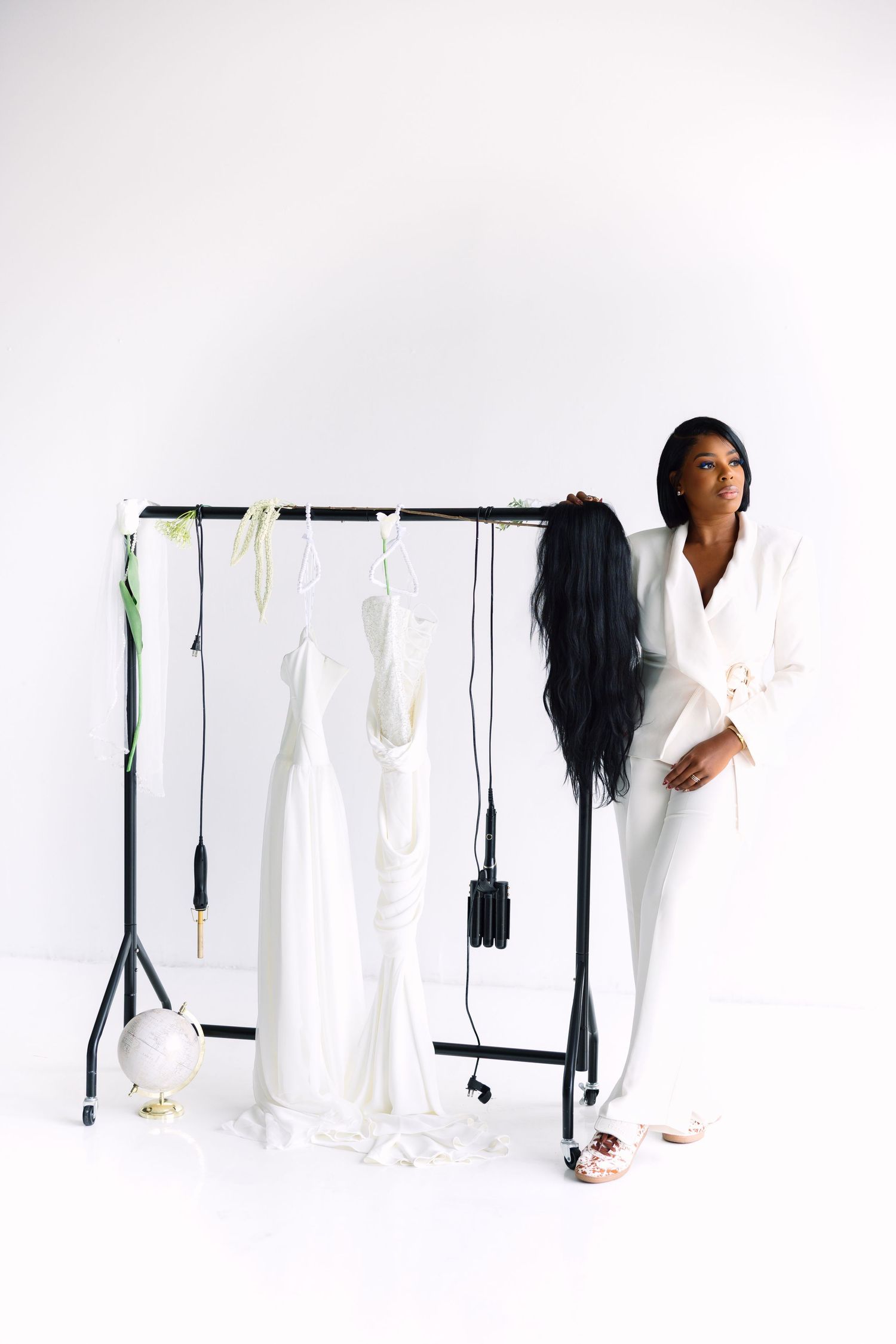 A stylish Black woman in an all-white suit leans against a clothing rack with elegant white garments hanging on it.