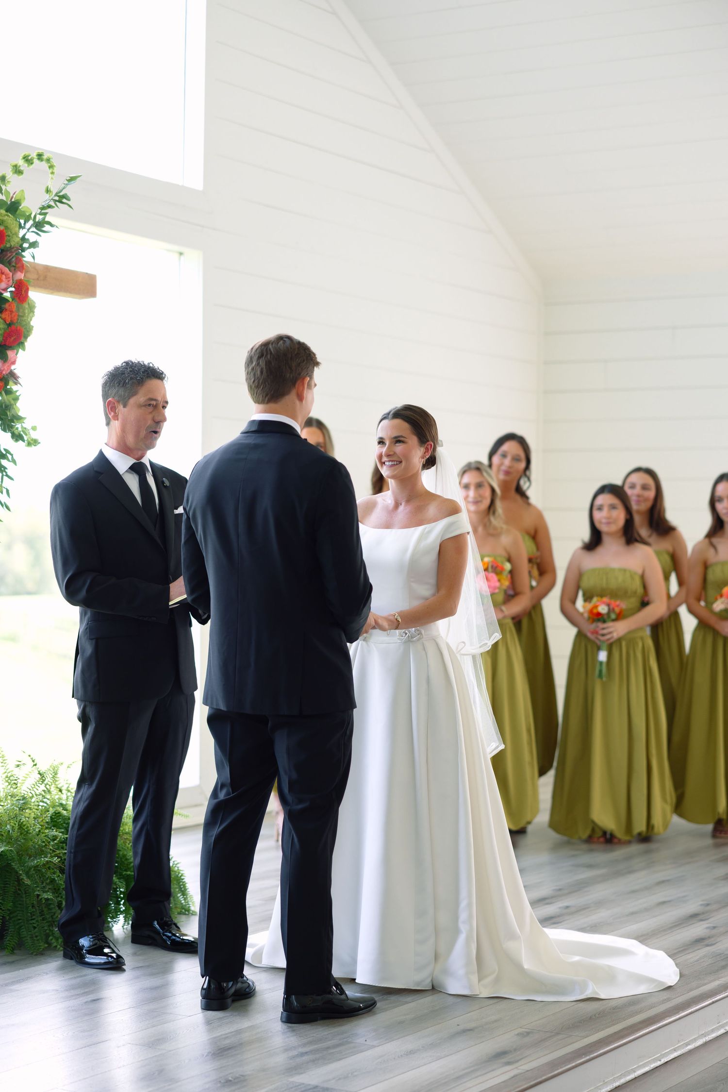 Bride smiling at groom during wedding ceremony with bridesmaids in olive green dresses standing behind her.