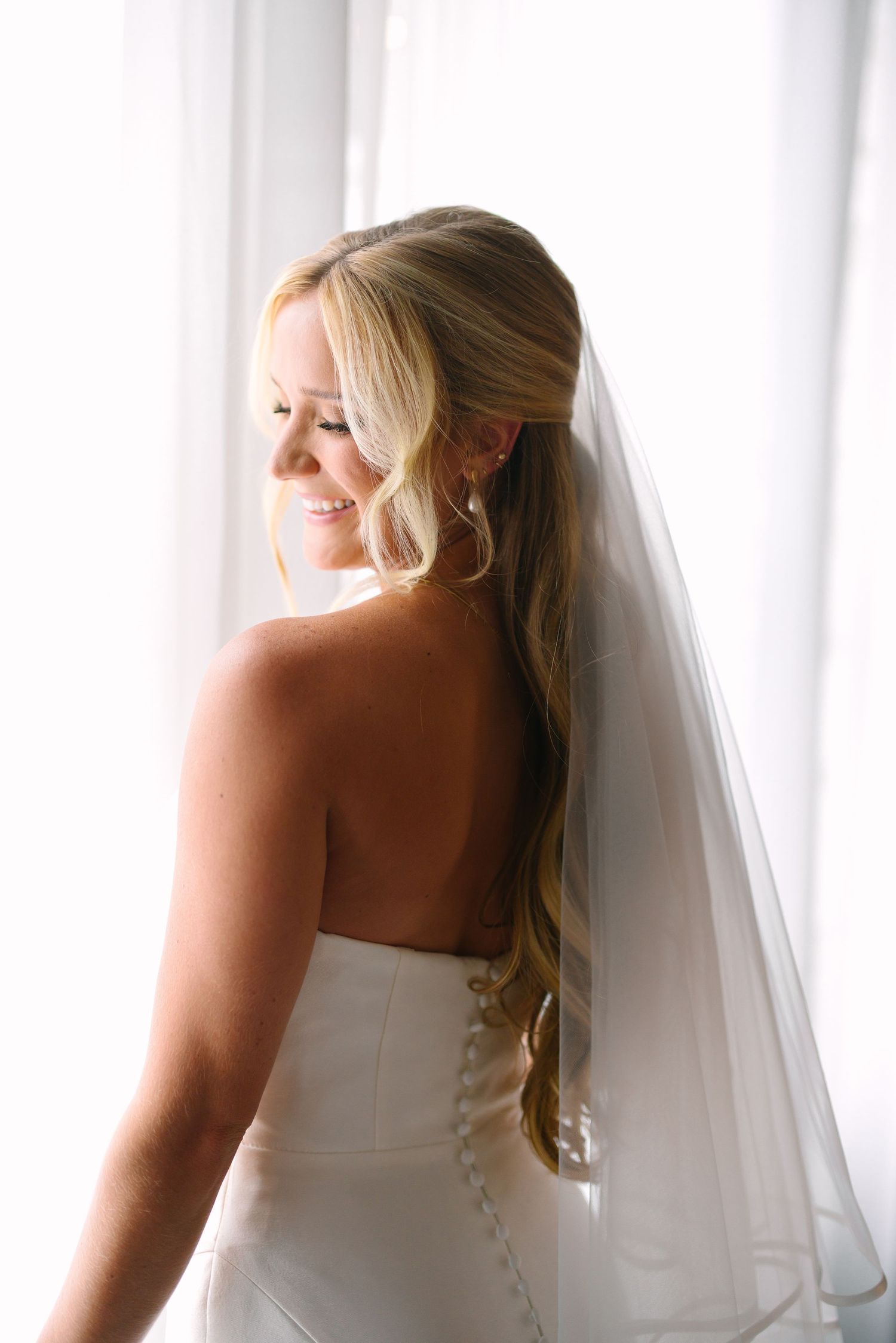 Back view of a bride in a strapless gown with flowing veil against white curtains.
