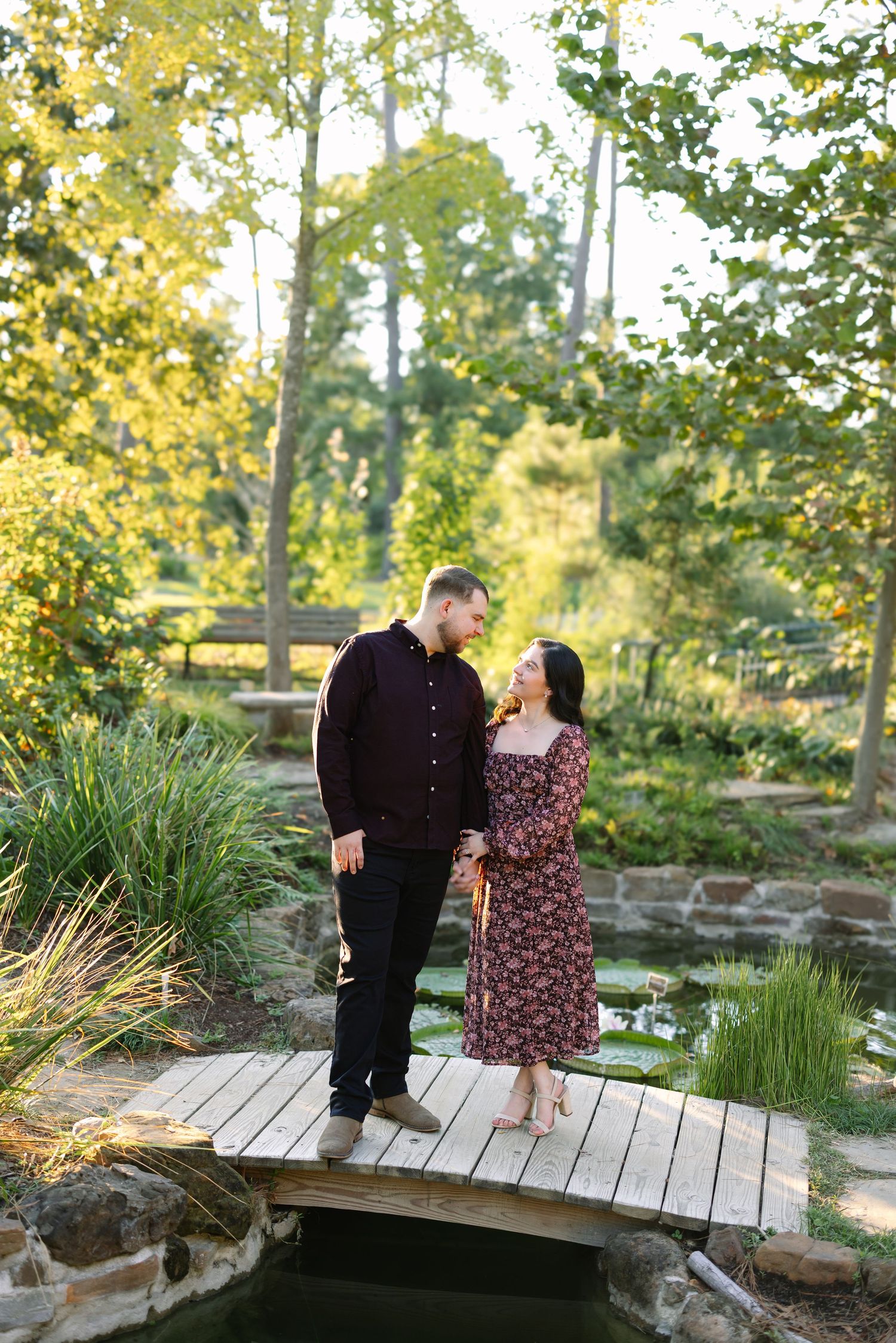 Couple in matching burgundy outfits standing on a wooden bridge surrounded by lush greenery and a pond.