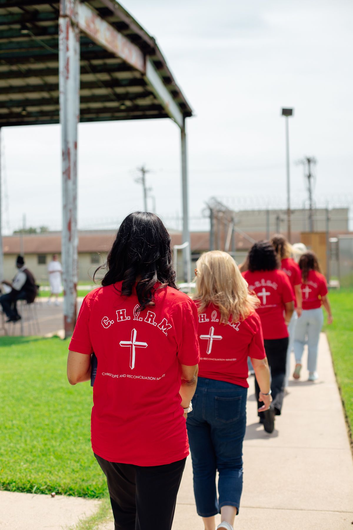 Prison ministry volunteers wearing red shirts with crosses walking along outdoor pathway.