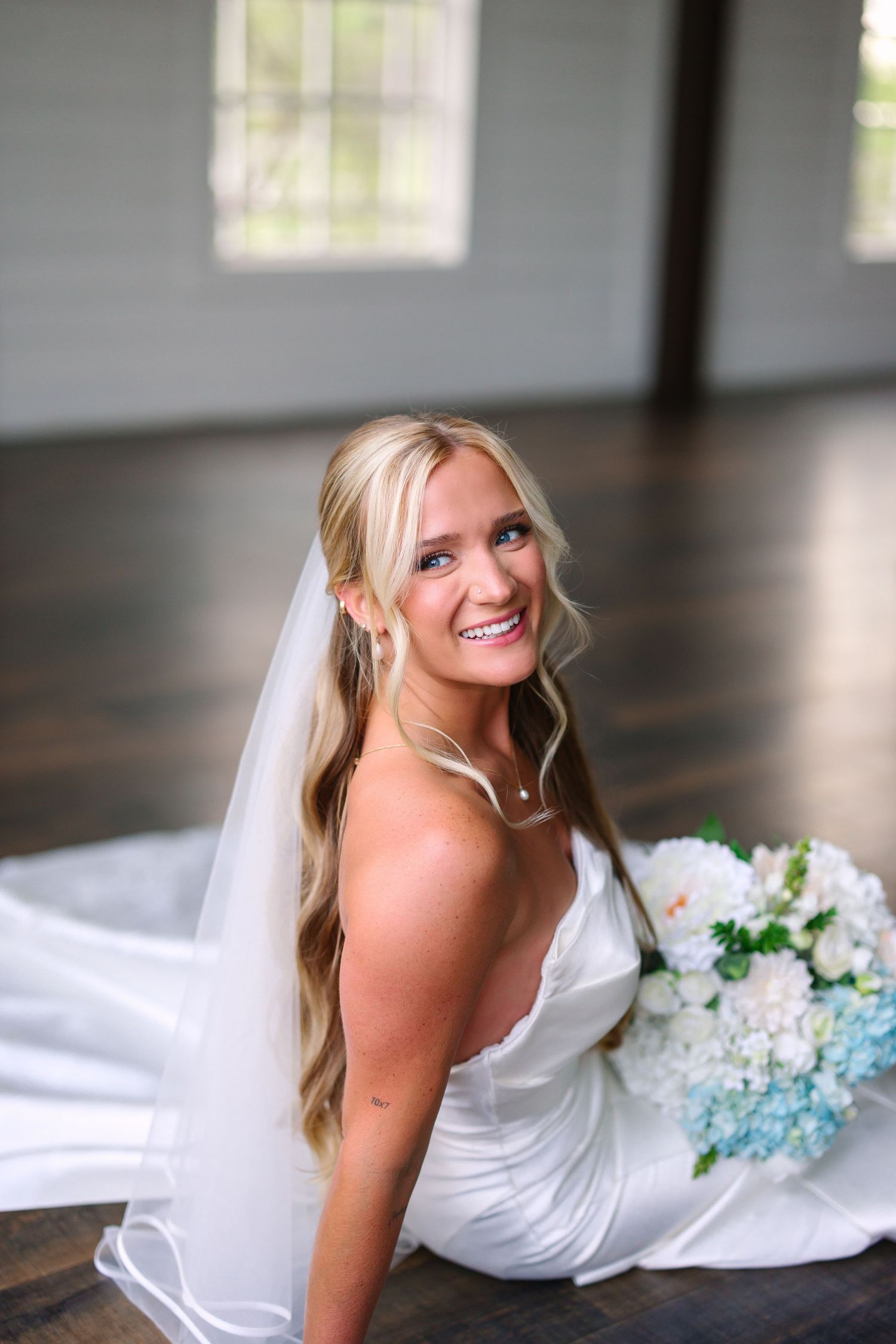 Bride in white wedding dress poses elegantly on floor with blue and white floral bouquet.