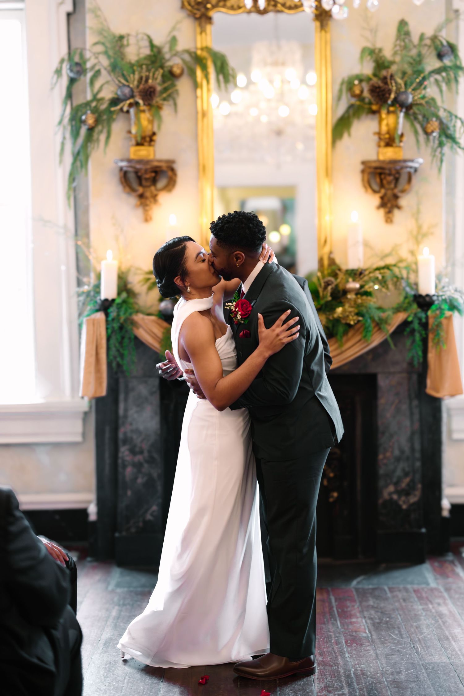 A bride and groom share their first kiss at the altar, surrounded by lush greenery and a golden fireplace mantle.