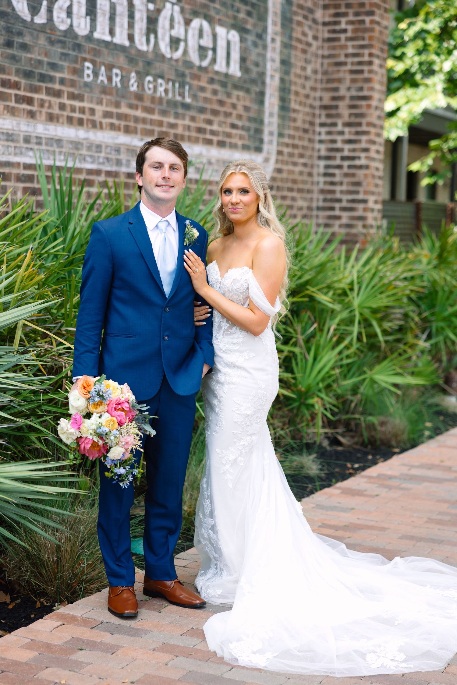 Bride in white gown and groom in blue suit pose together outside Canteen Bar and Grill on their wedding day.