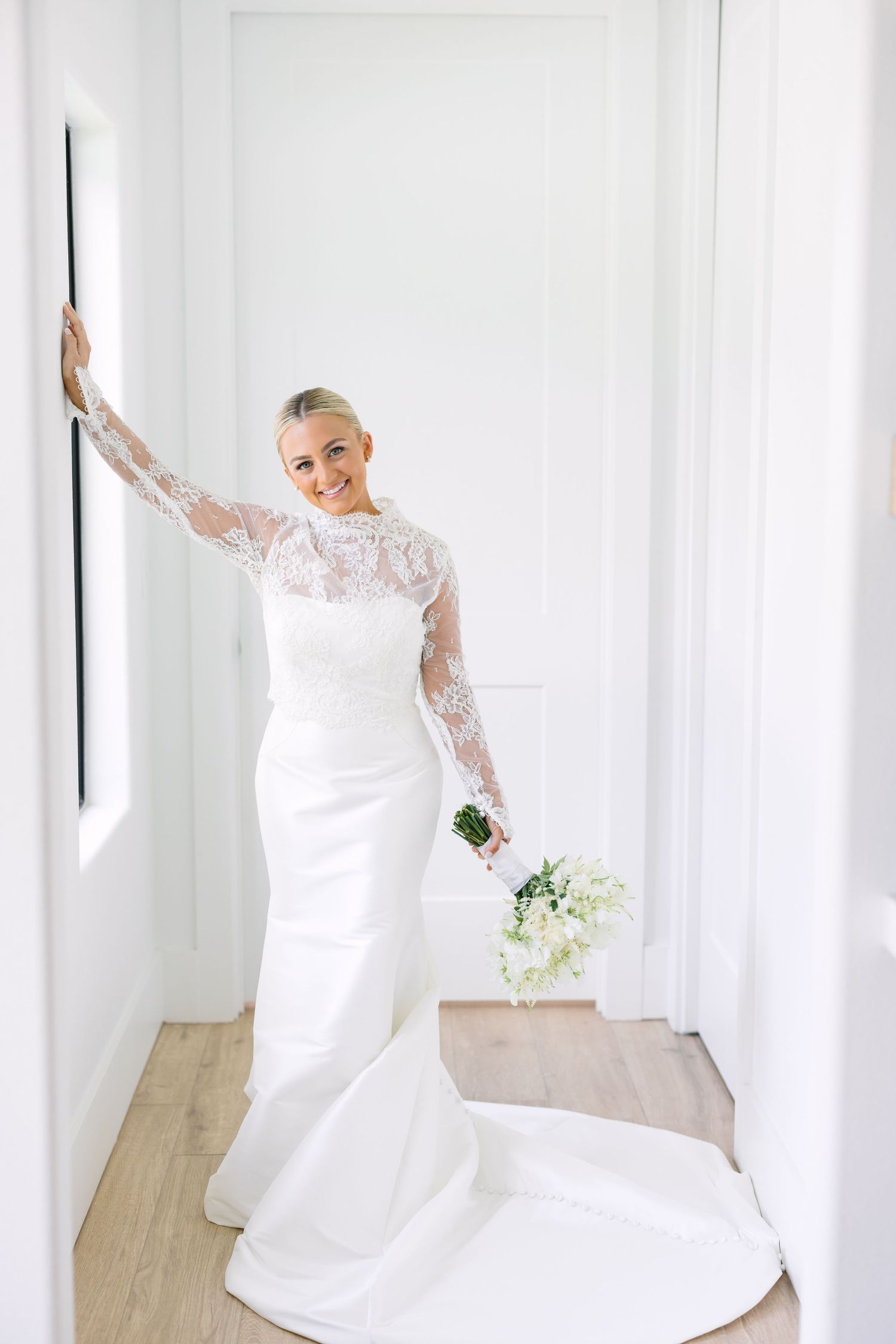 Elegant bride in long-sleeve lace wedding gown holds white bouquet while posing in a bright white hallway.