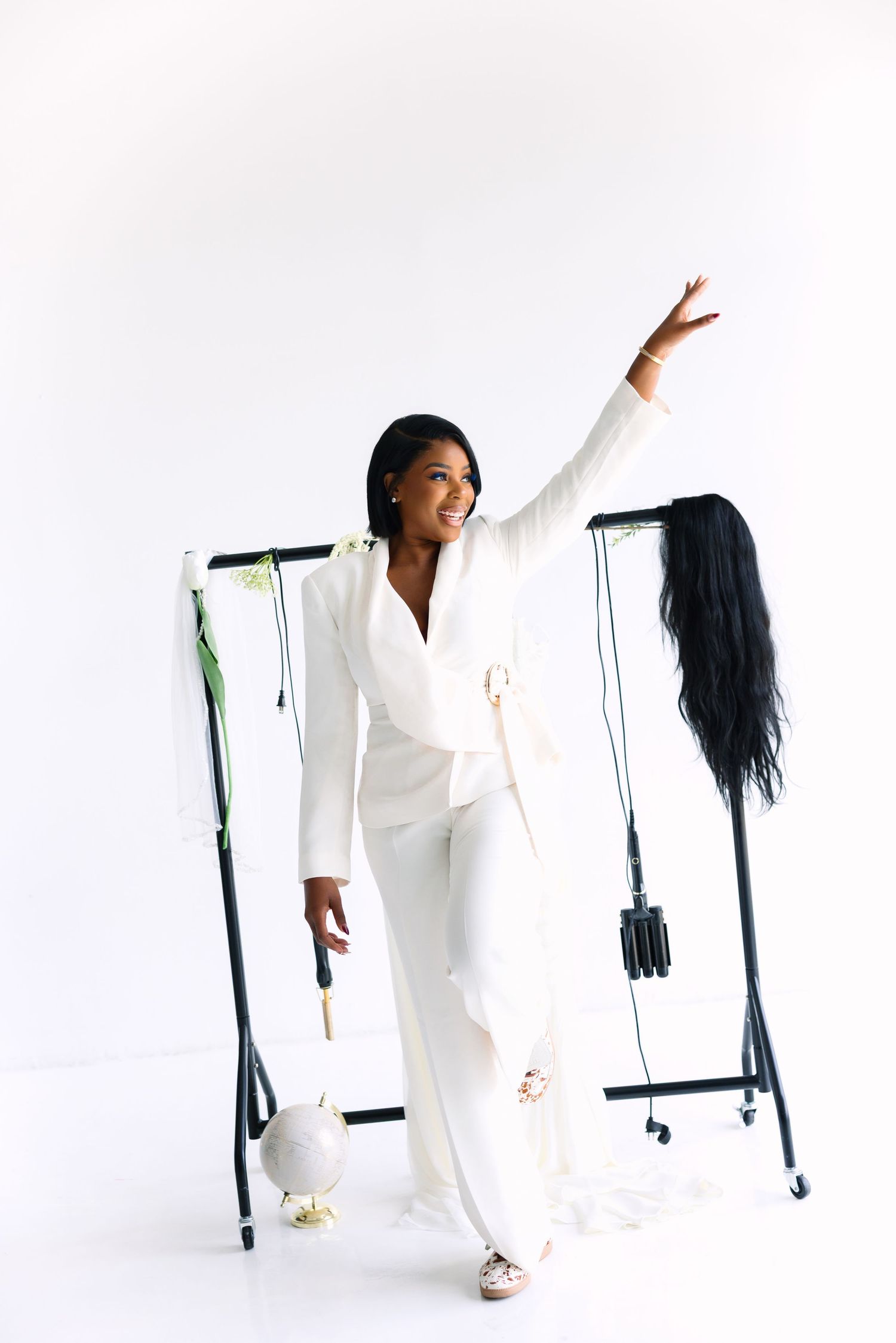 Woman in white pantsuit poses confidently with raised arm beside a clothing rack on a bright white background.