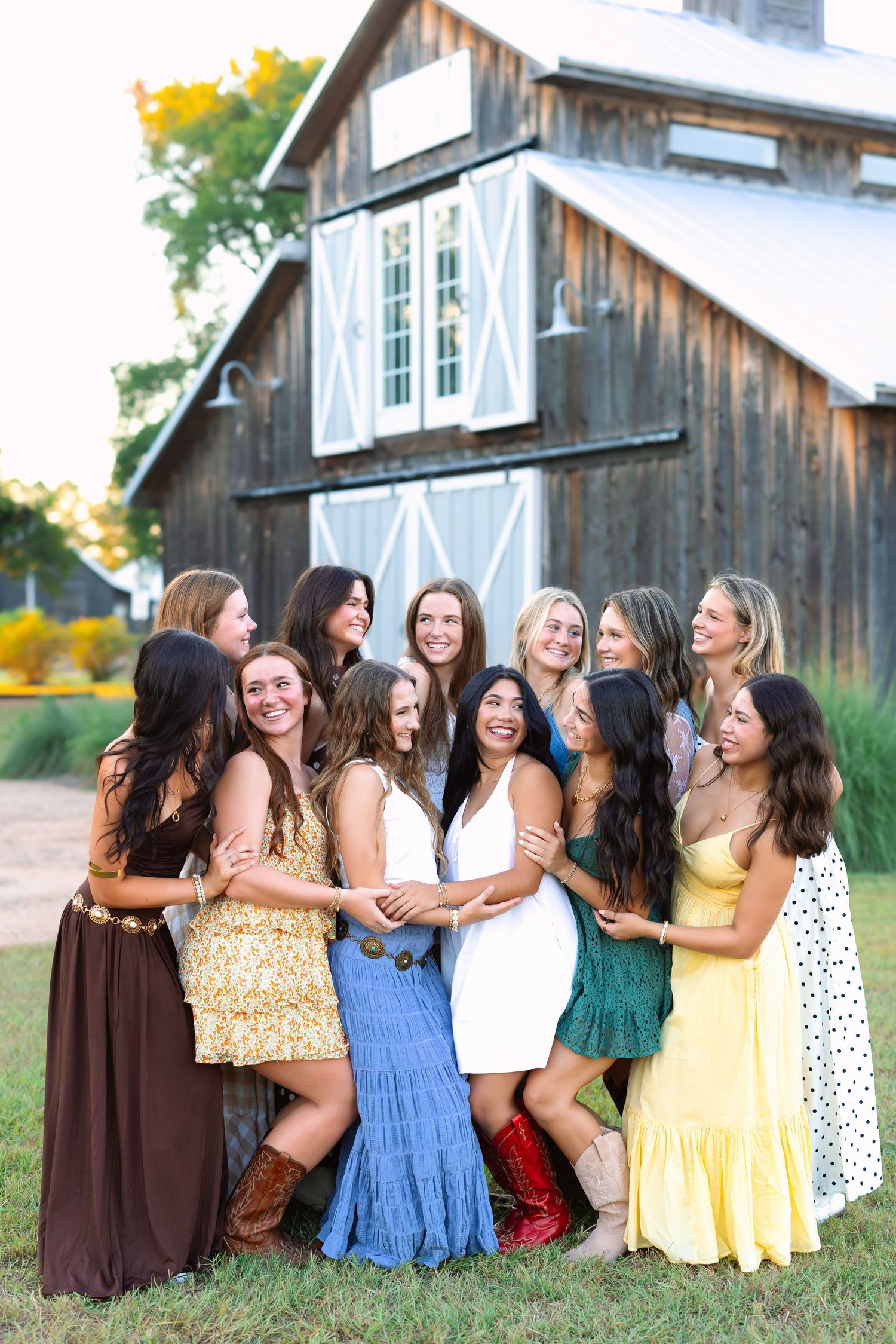 Group in sundresses and boots sharing a candid moment of laughter by a rustic barn.