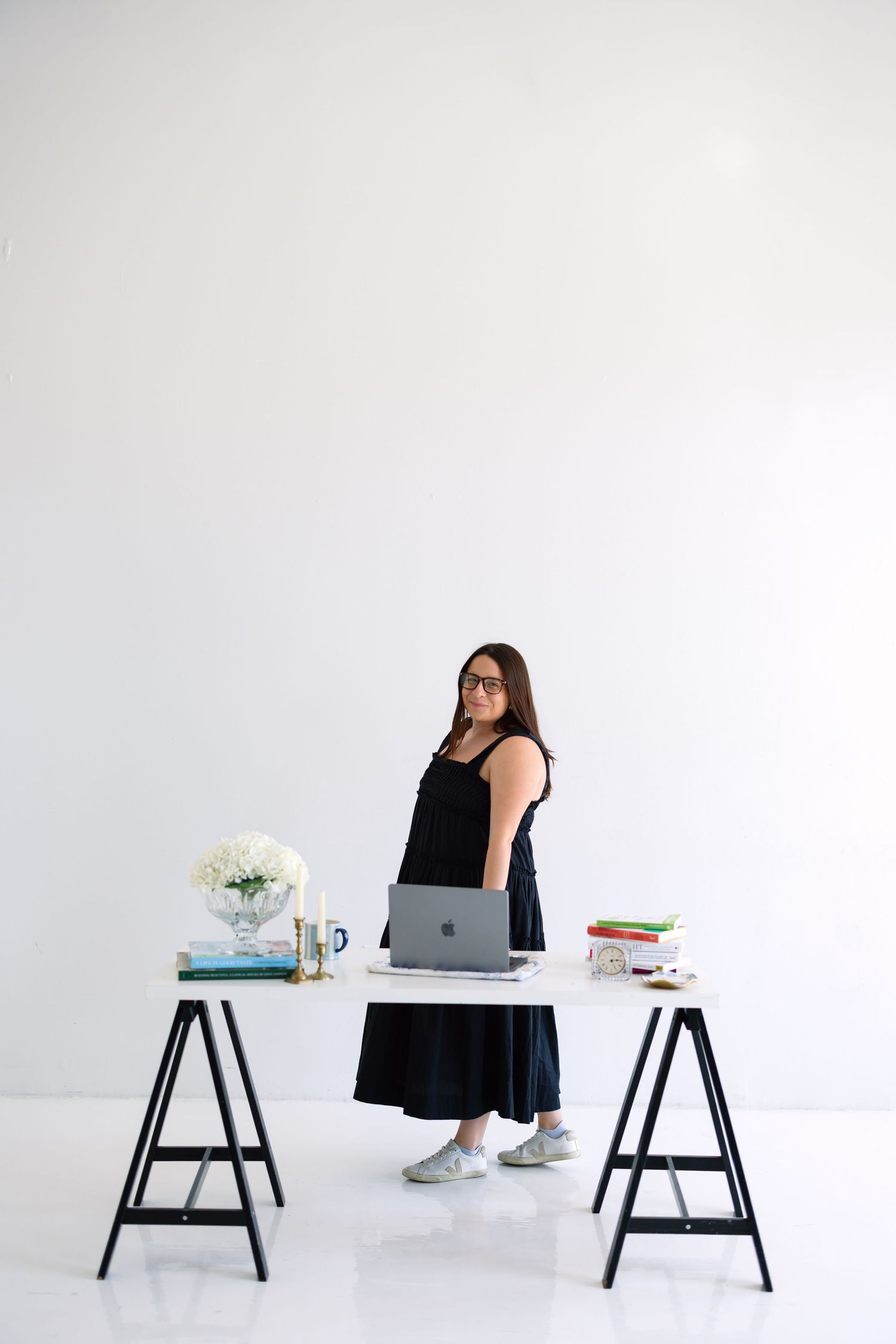 Woman in black dress standing at a white trestle desk with laptop, flowers, and books in a bright white studio.