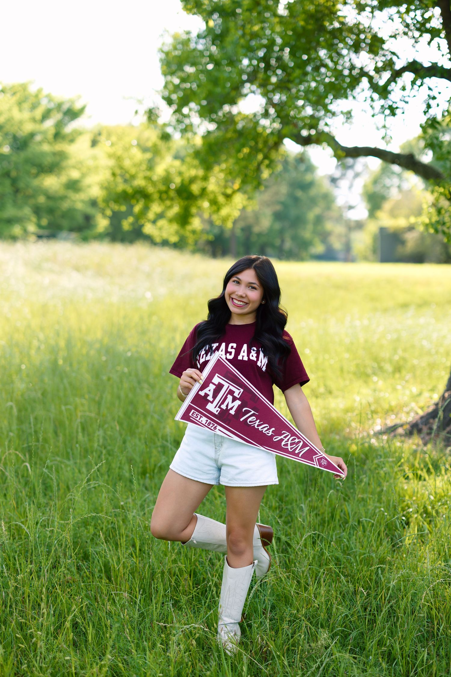 Smiling young woman holding a Texas A&M pennant flag in a green field wearing a maroon Aggies t-shirt and white shorts.