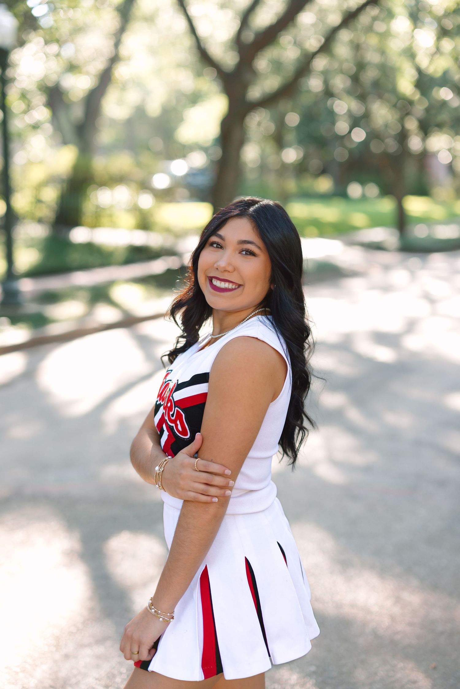 Woman in white cheerleader uniform poses outdoors under trees with dappled sunlight.