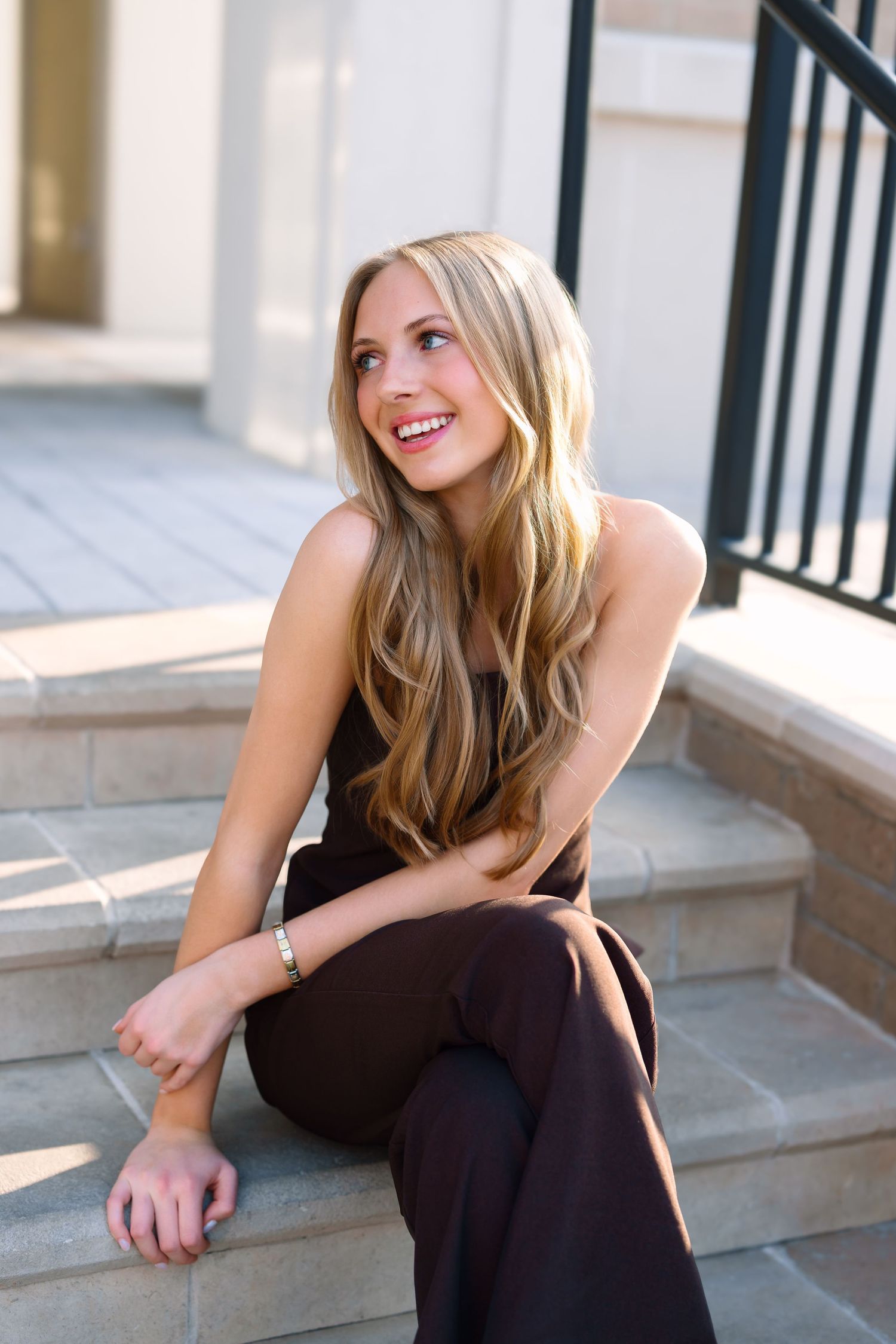 Smiling young blonde woman in dark jumpsuit sitting on outdoor stone steps in sunlight.
