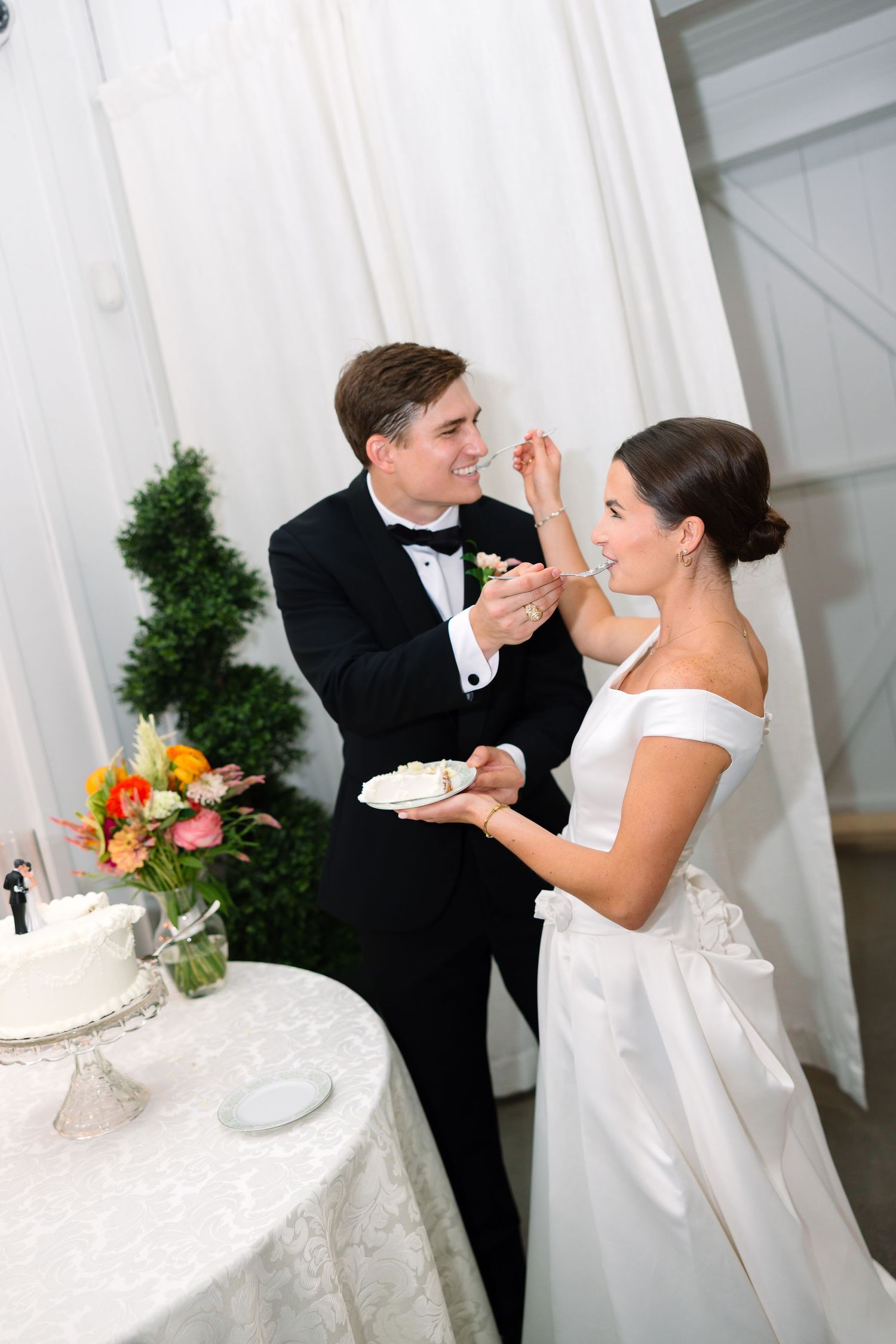 Bride and groom sharing wedding cake at reception, groom feeding bride in elegant white venue.