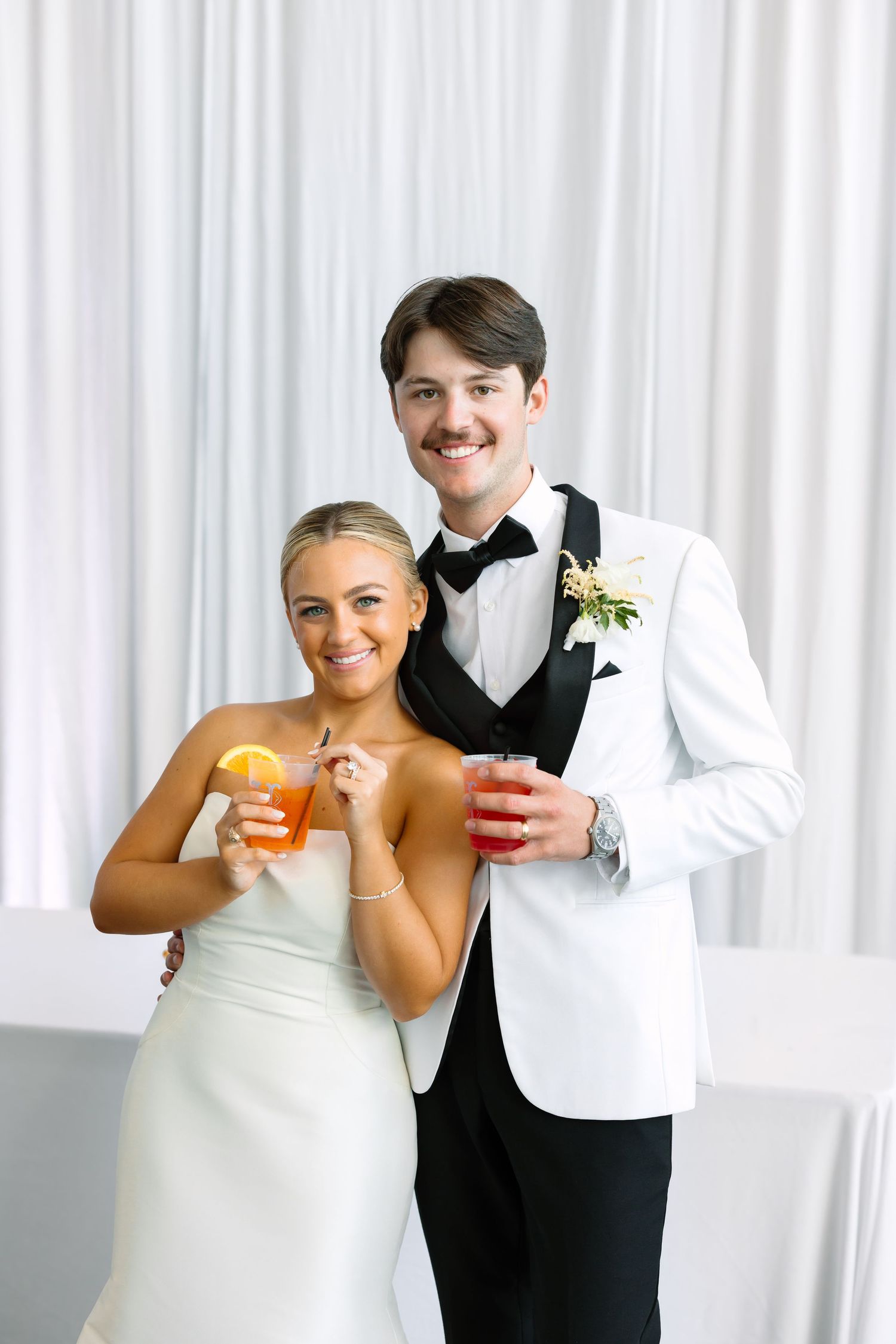 Bride and groom smiling together holding colorful cocktails, dressed in wedding attire with white drape backdrop.