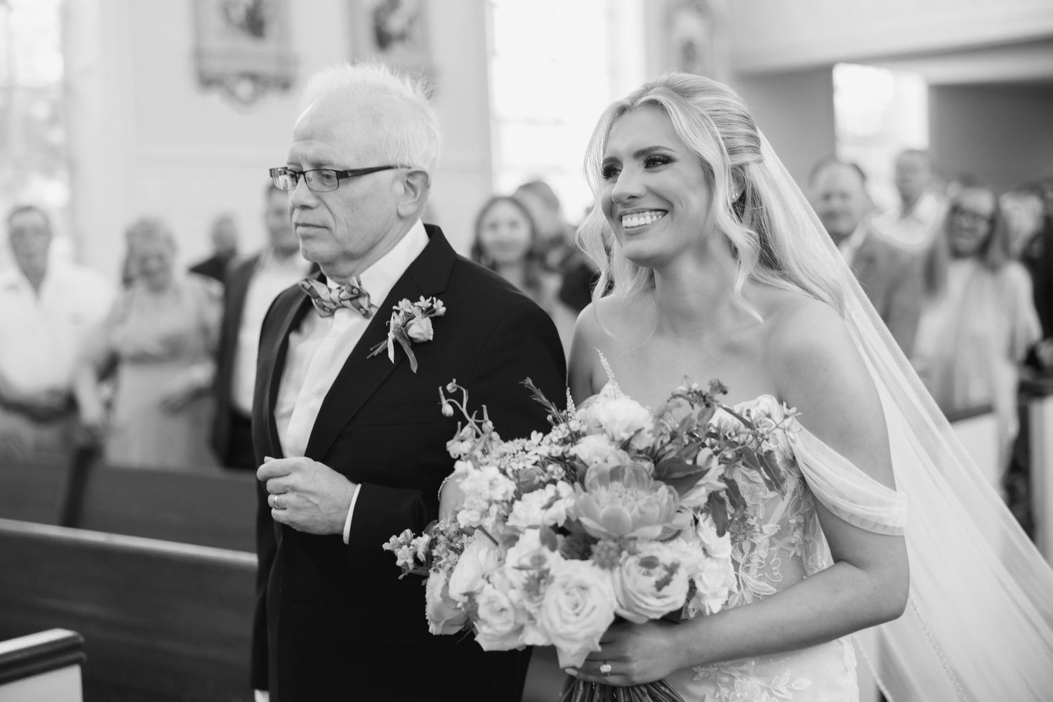 Bride walks down church aisle with older man in tuxedo, holding floral bouquet, in black and white wedding photo.