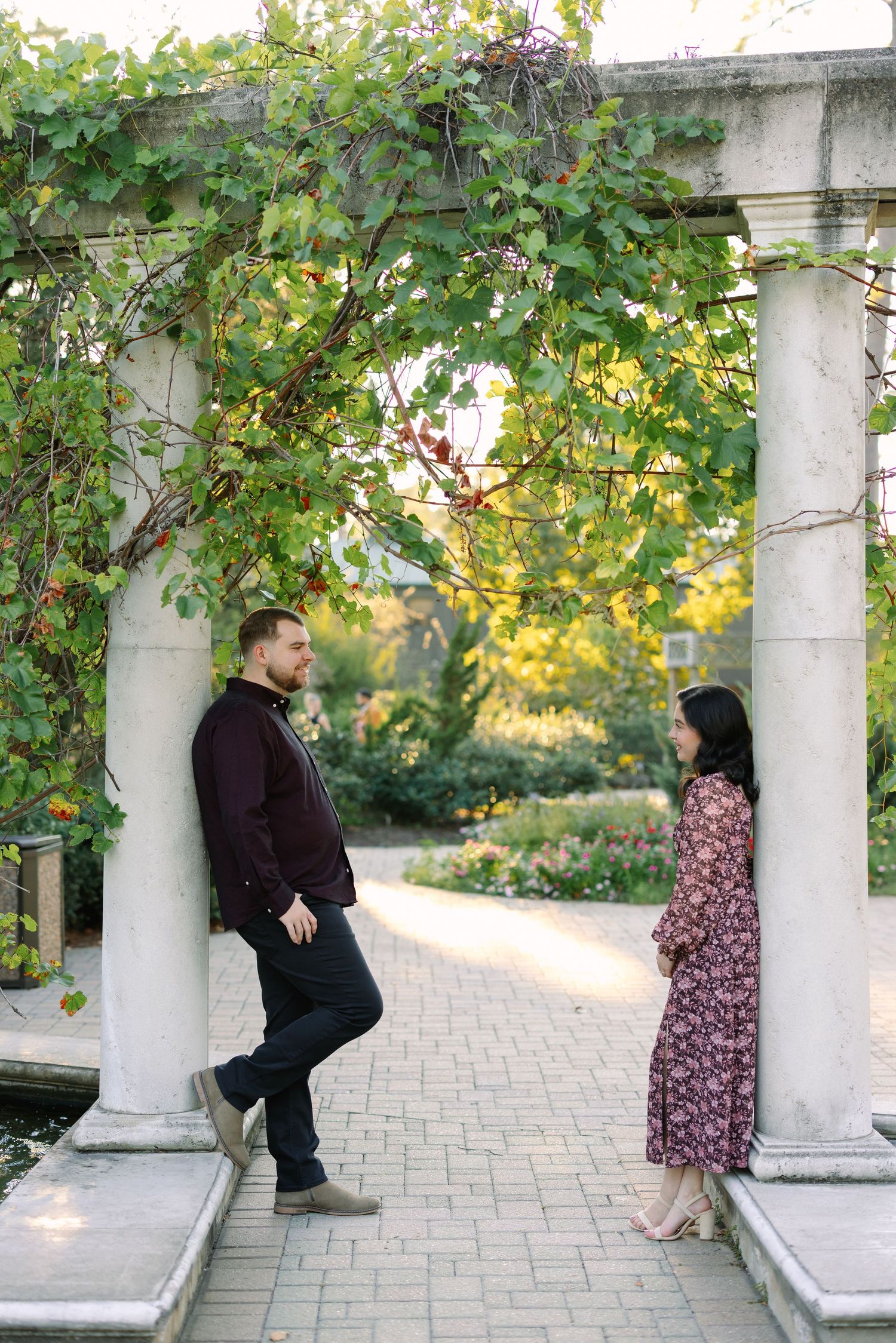 Couple sharing a romantic moment under a vine-covered pergola with white columns in a lush garden setting.