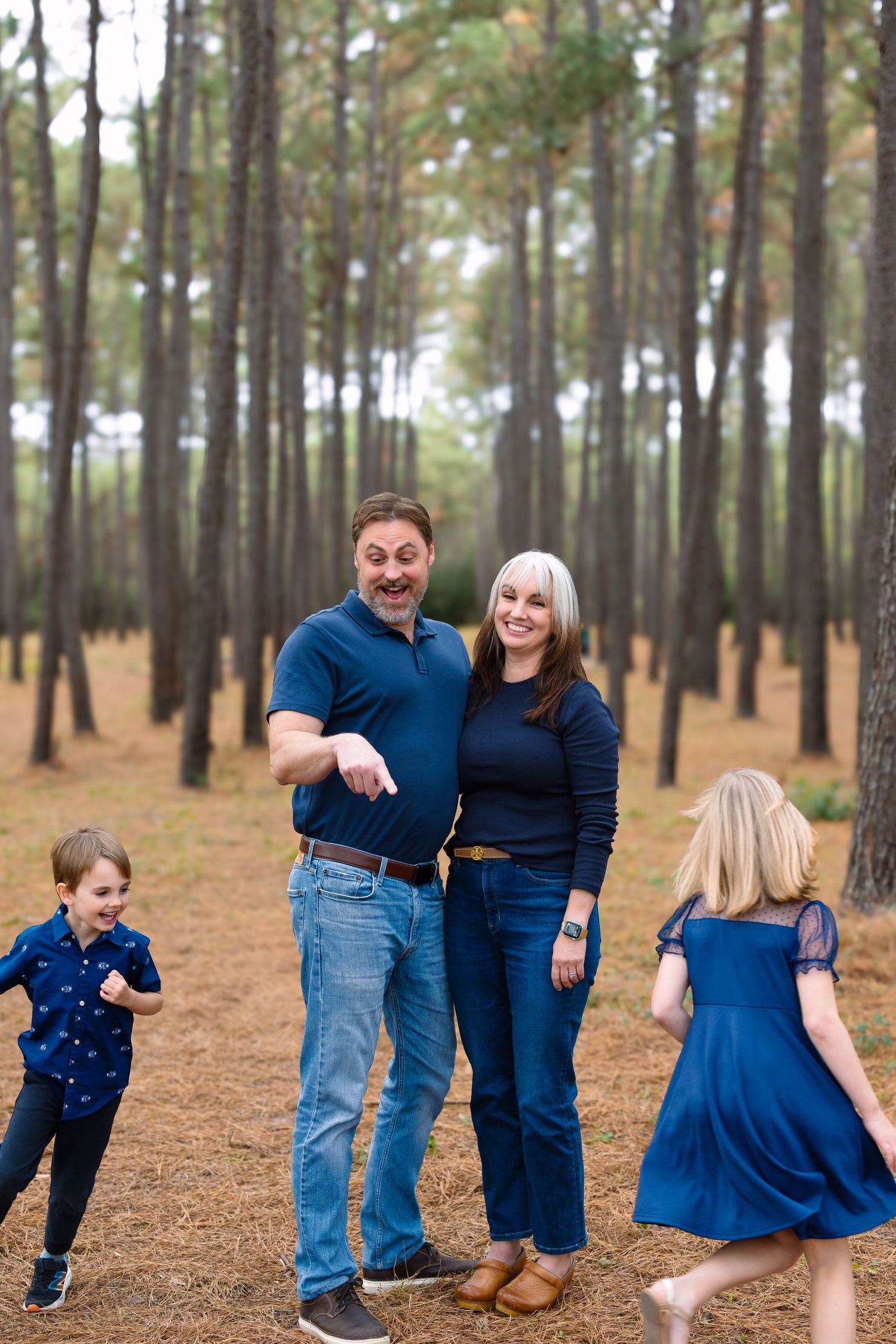 Family in coordinating blue outfits laughing together in a pine forest, with two children running toward their parents.
