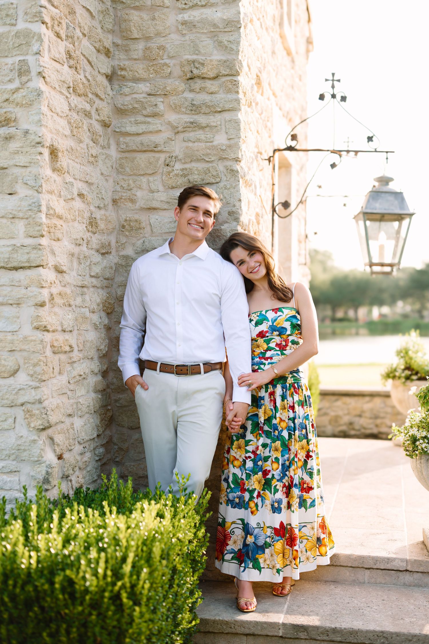 A smiling couple poses by a stone building, the woman in a floral dress leaning on a man in a white shirt and khakis.