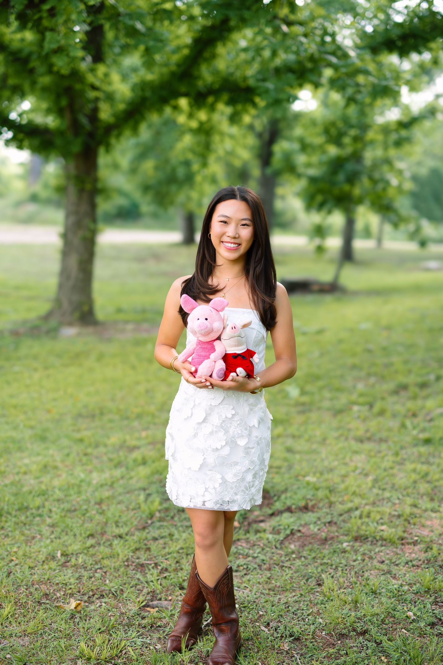 Smiling young woman in white dress and cowboy boots holding stuffed pig toys in a green park.