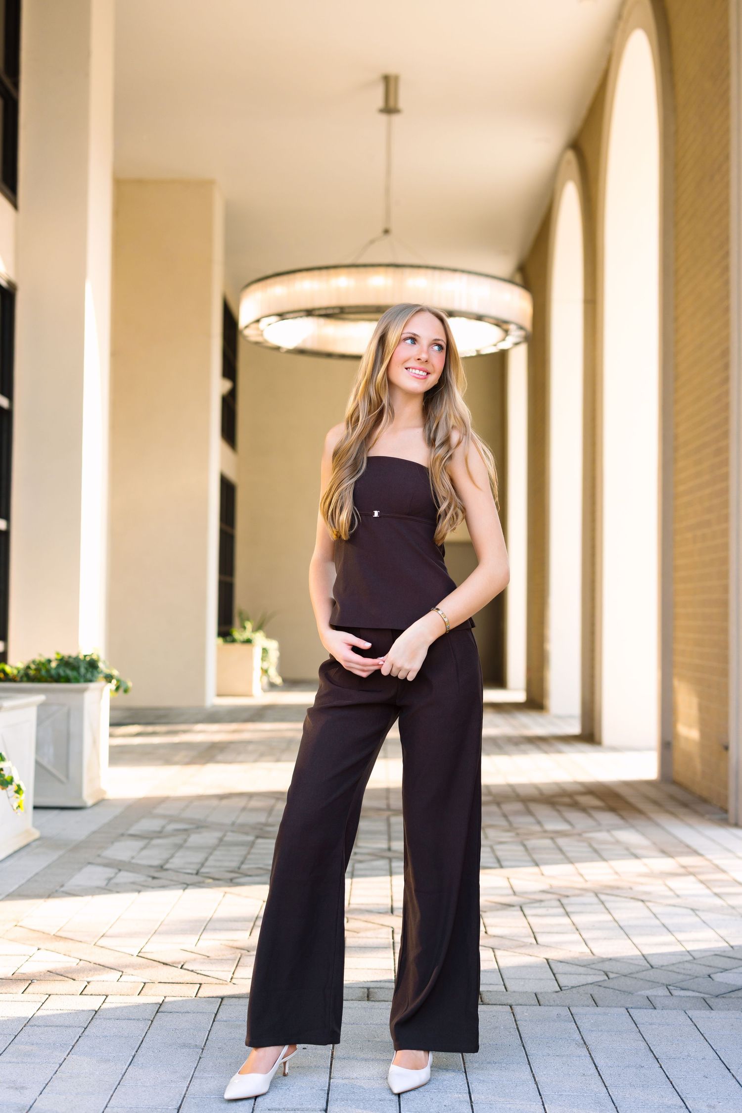 Pregnant woman in black strapless jumpsuit and white heels smiling in elegant architectural hallway.