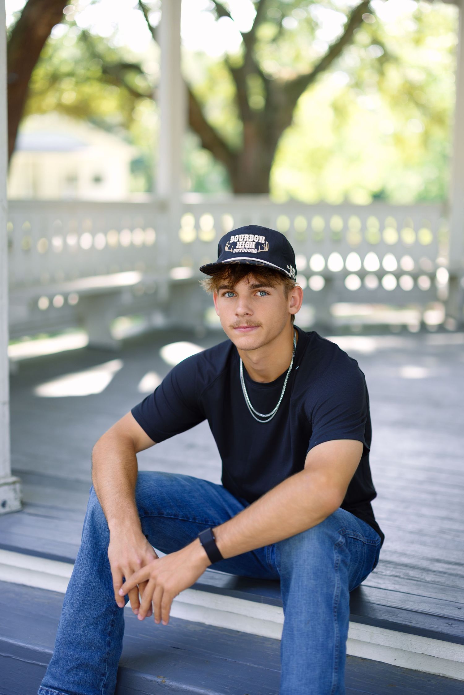 Young man in navy cap, black t-shirt, and jeans sits on steps of a white gazebo surrounded by trees.