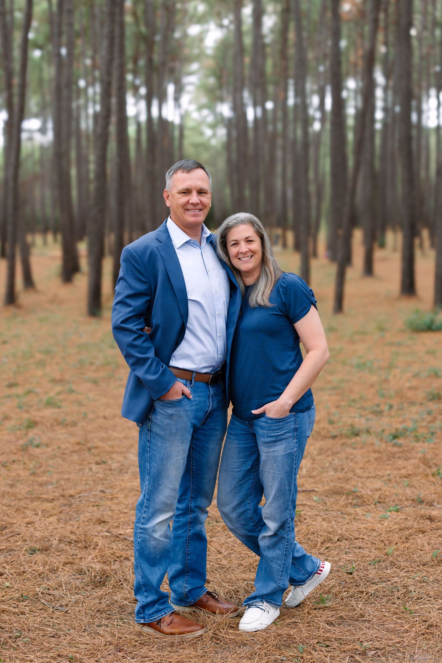 Smiling middle-aged couple in matching blue outfits posing together in a pine forest with needle-covered ground.