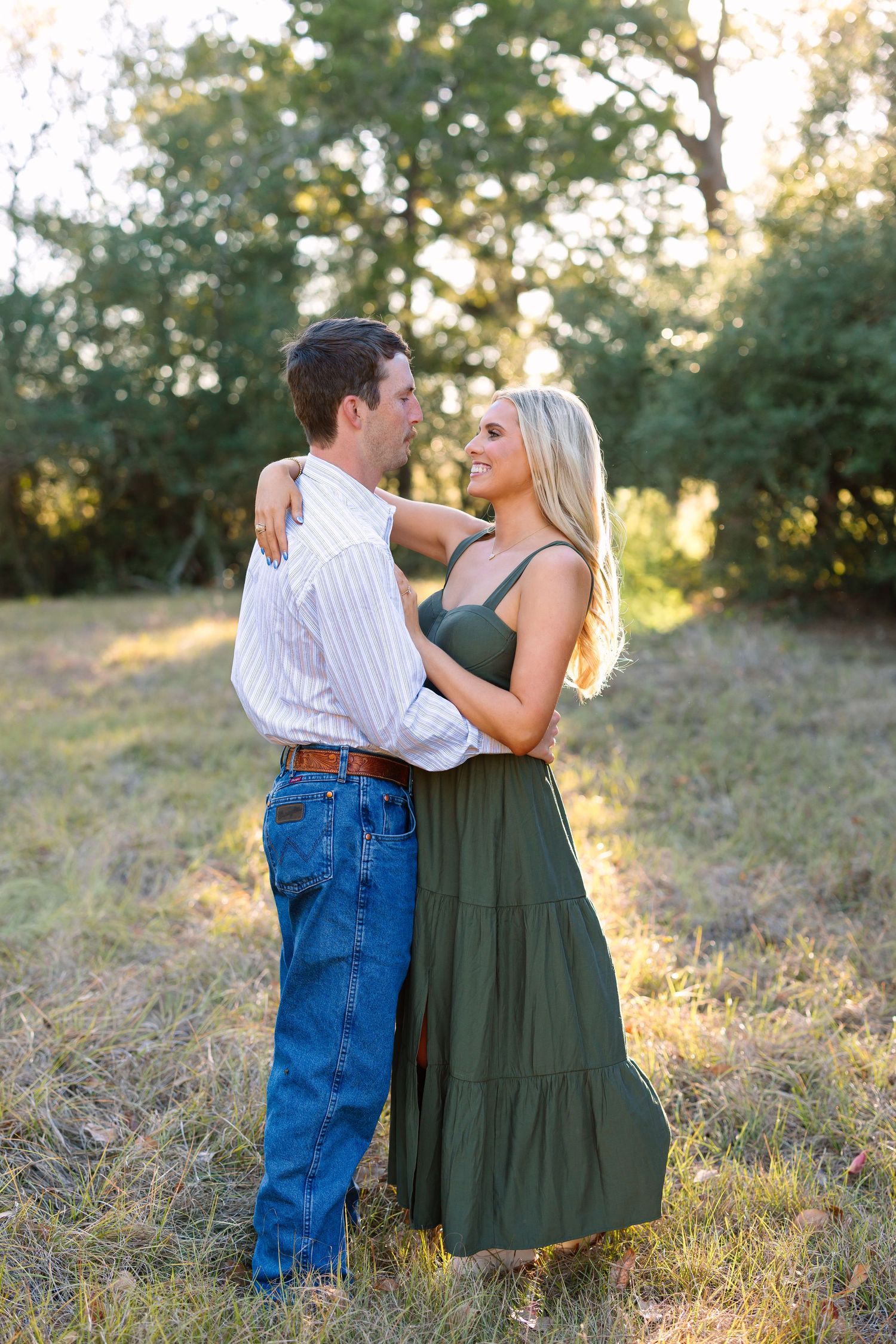 Couple embracing in a sunlit field, woman in green maxi dress smiling at man in striped shirt and jeans.