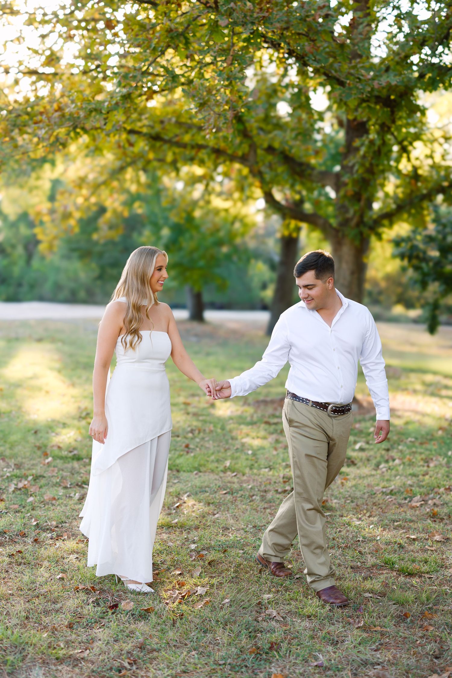 Couple holding hands and walking through a sunlit park, woman in white dress and man in white shirt and khaki pants.