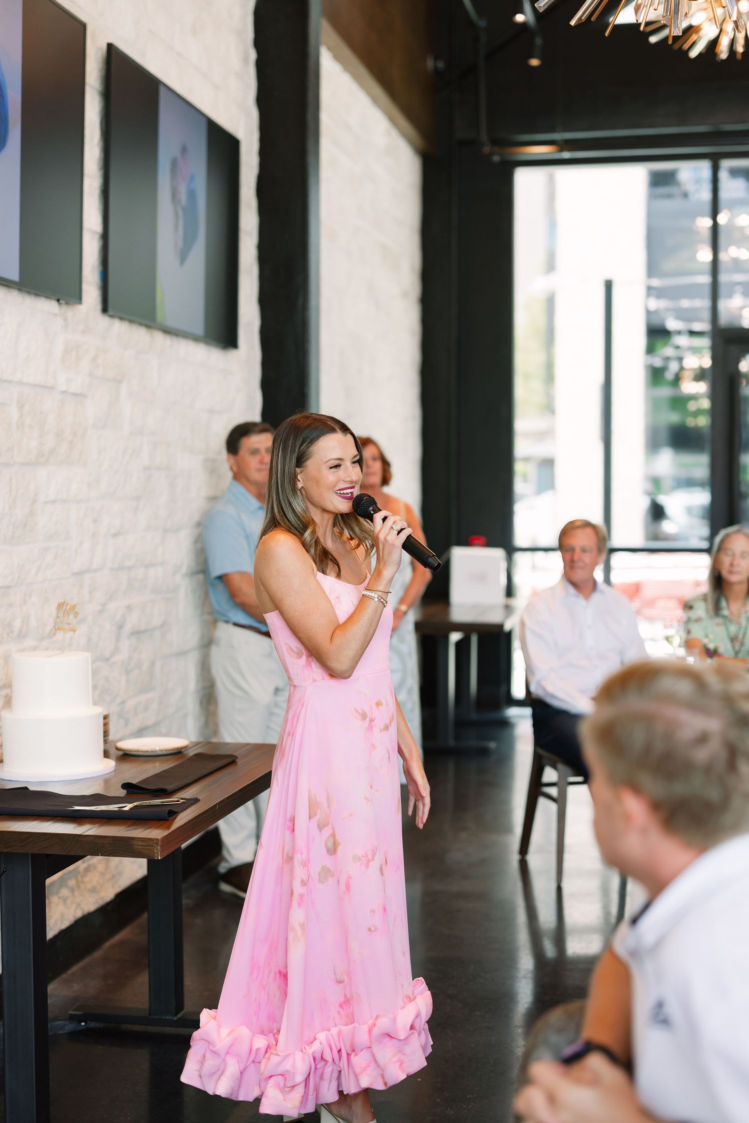 Woman in pink floral ruffle dress speaks into microphone at elegant indoor event with guests seated nearby.