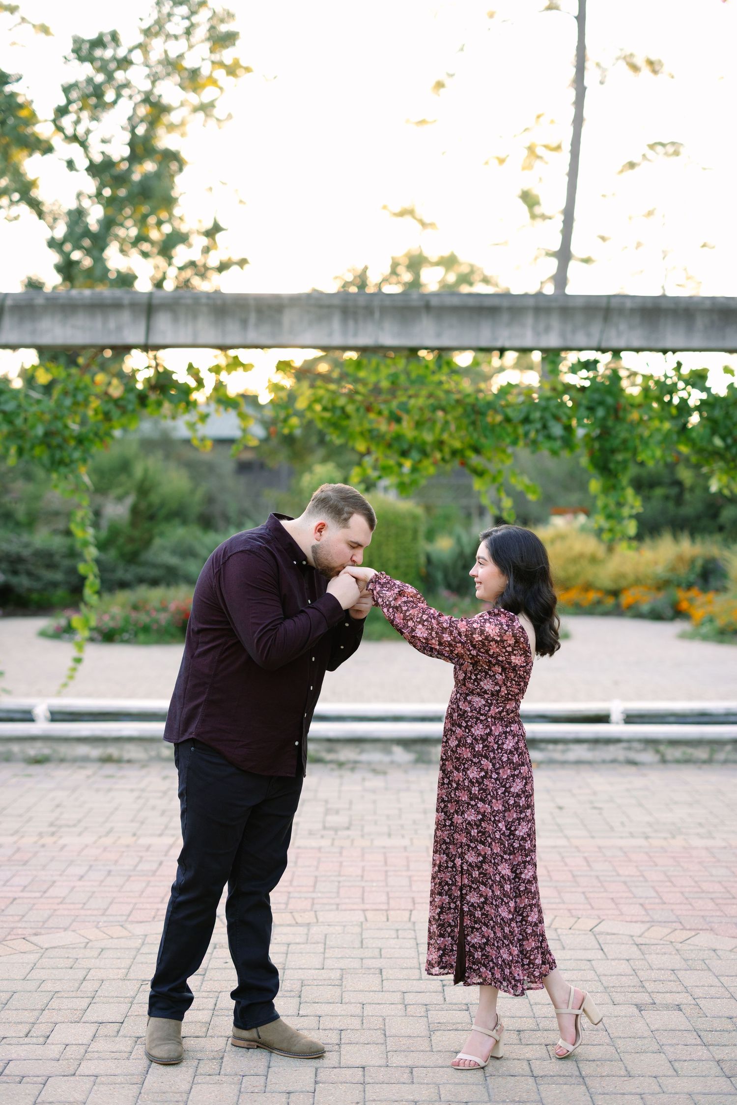 A man in a burgundy shirt kisses a woman's hand in a garden, as she smiles in a floral dress.