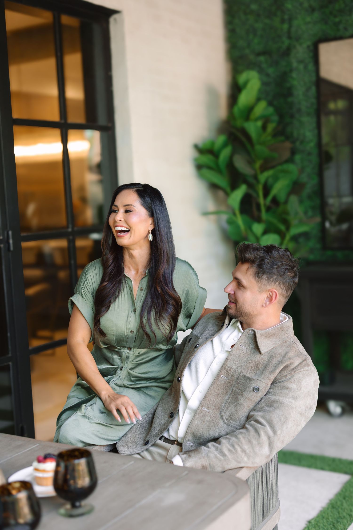 A laughing couple in green and grey outfits enjoy a relaxed outdoor dining moment together.